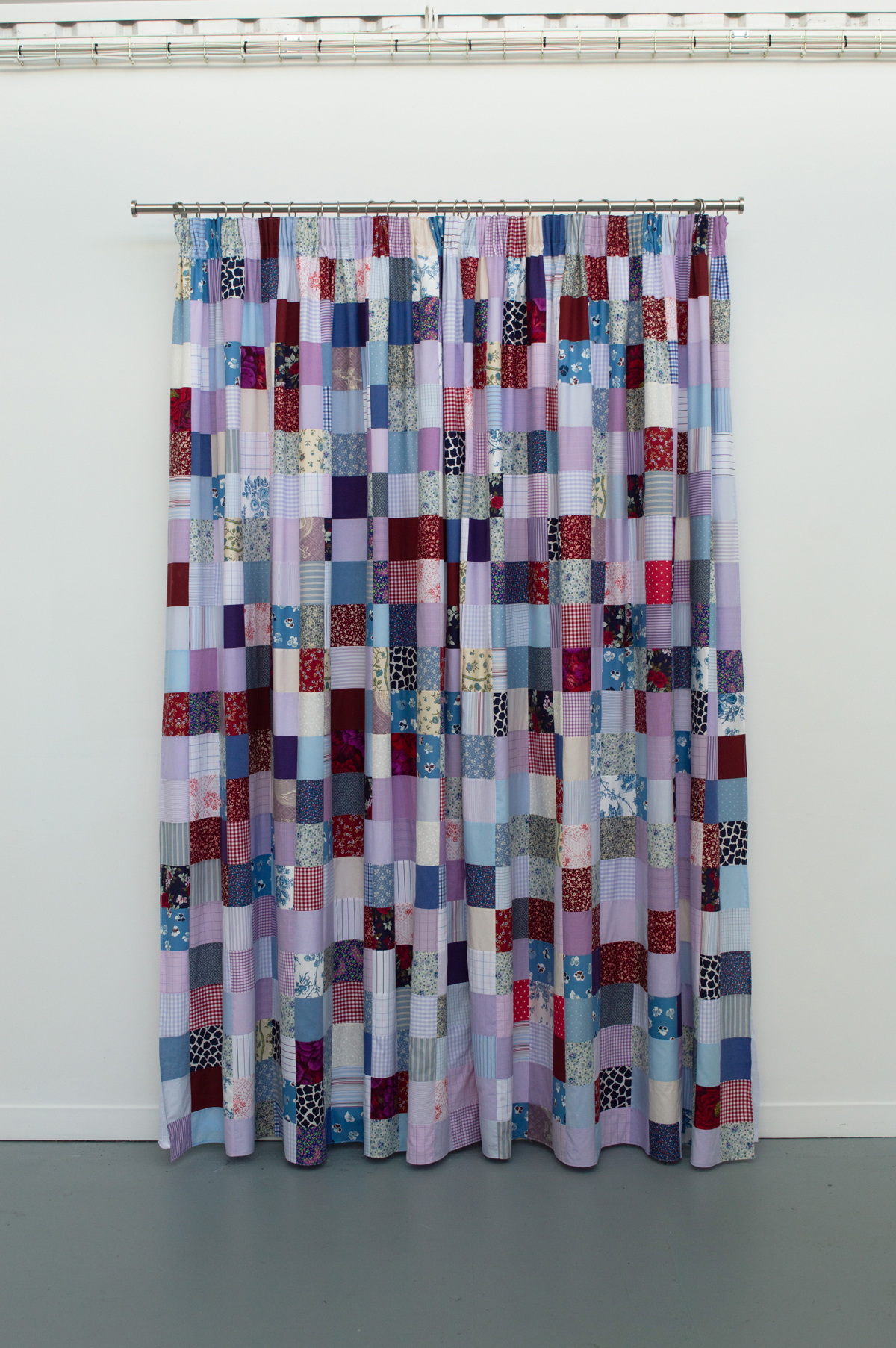 Patchwork curtain with rectangular fabric squares in blues, reds, purples, and whites hung on metal rail against white wall.