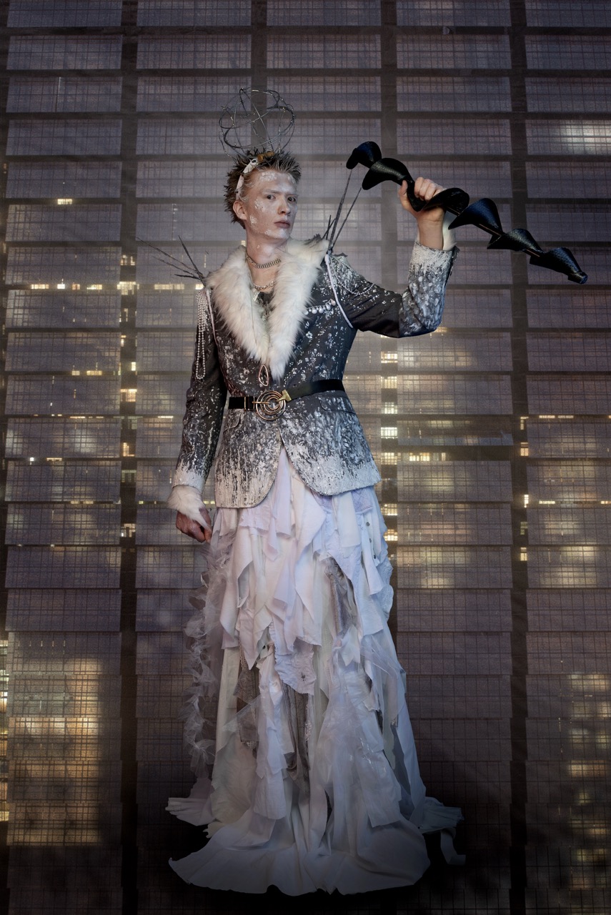 A model wearing a silver textured jacket and layered white skirt holds a black sculptural accessory, standing before a woven screen backdrop.