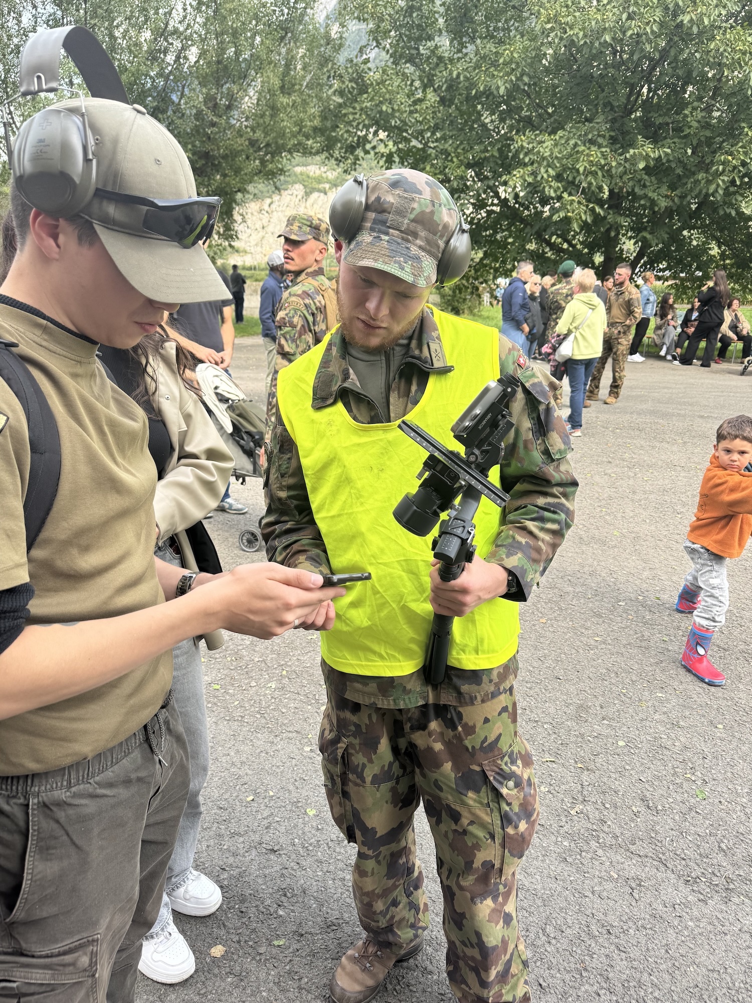 Two soldiers in camouflage uniforms and caps examining equipment, one wearing bright yellow high-vis vest, with crowd and trees in background.