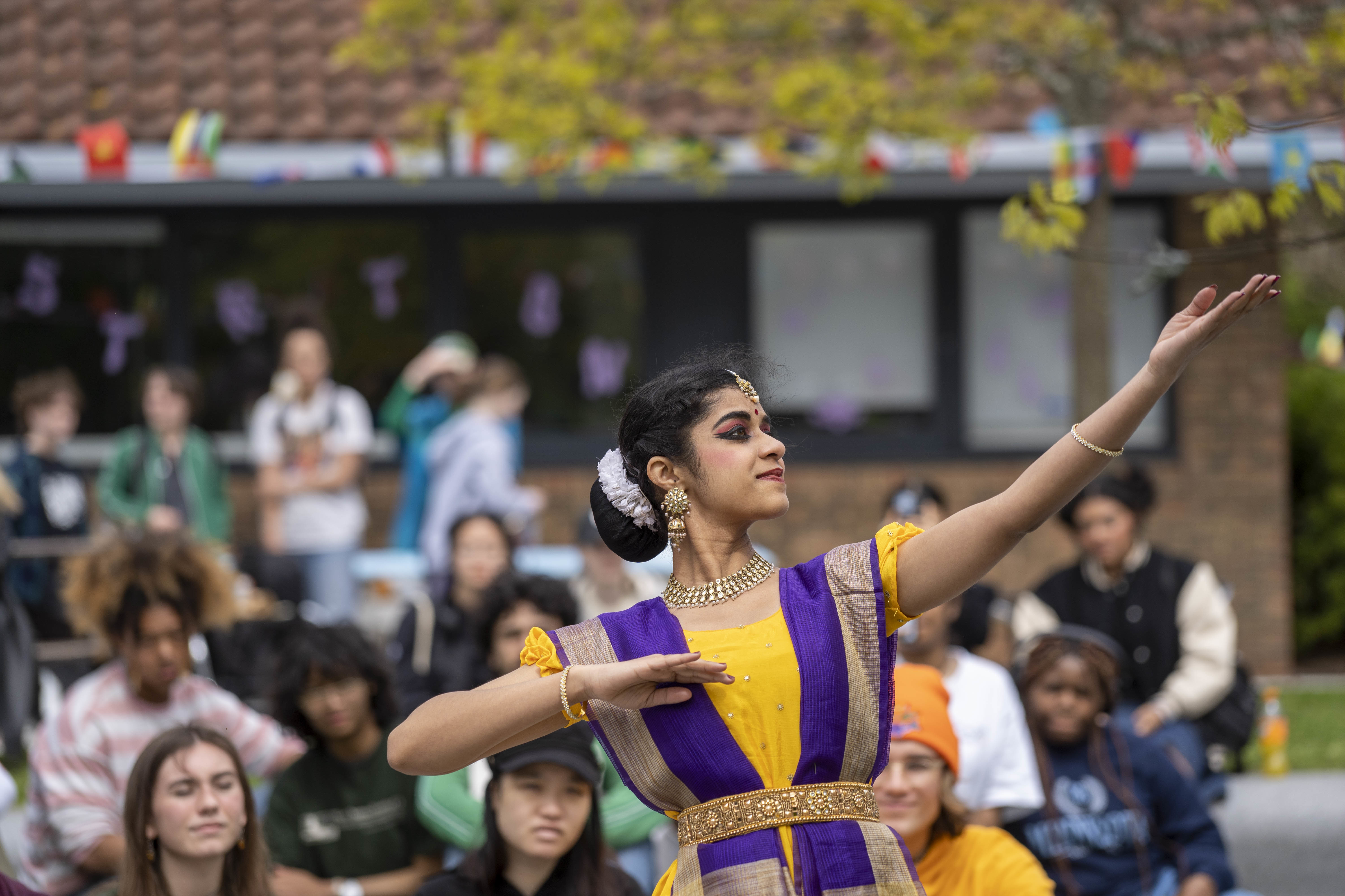 A person dressed in traditional Indian costume dancing. AUB students gather outside on campus to watch.