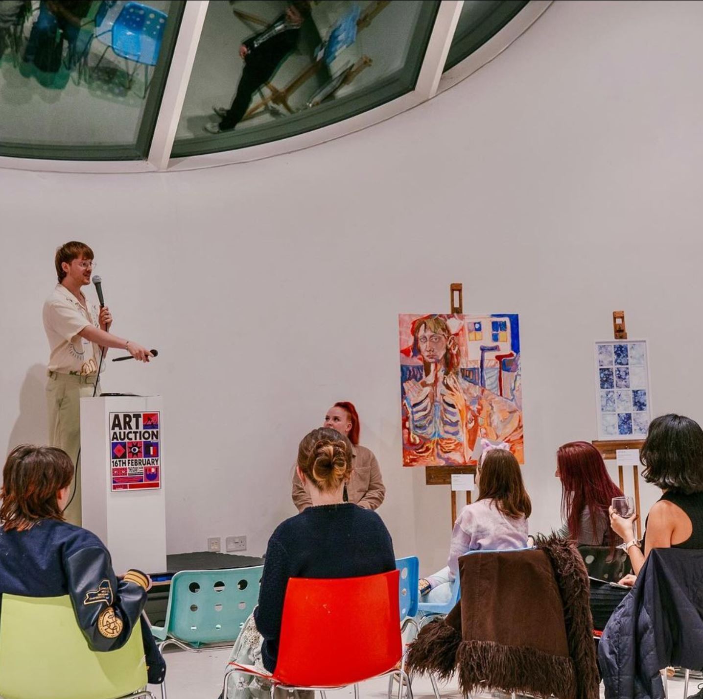 Man presenting to seated audience in white gallery space with colourful artwork displayed on walls and curved glass ceiling above.
