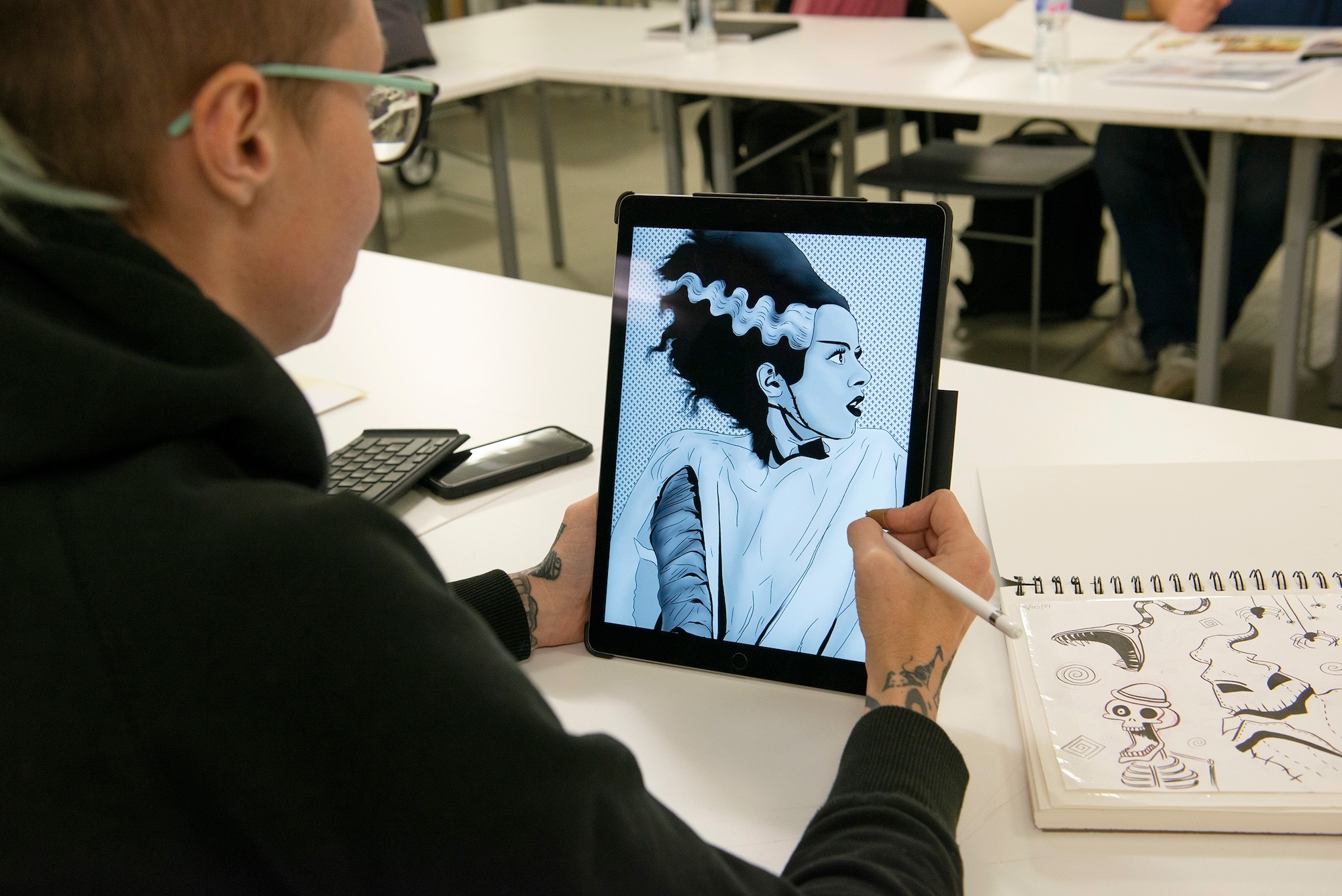 Person with glasses drawing on tablet displaying blue-tinted anime character, sketchbook and laptop on white table
