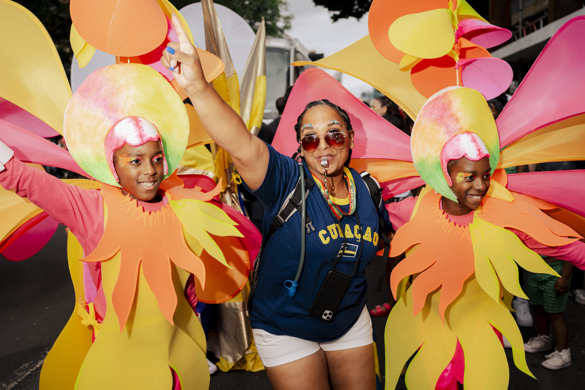 Three carnival performers, two in bright and colourful abstract costumes, the other in shorts and a blue top with "Curaçao" written on the front.