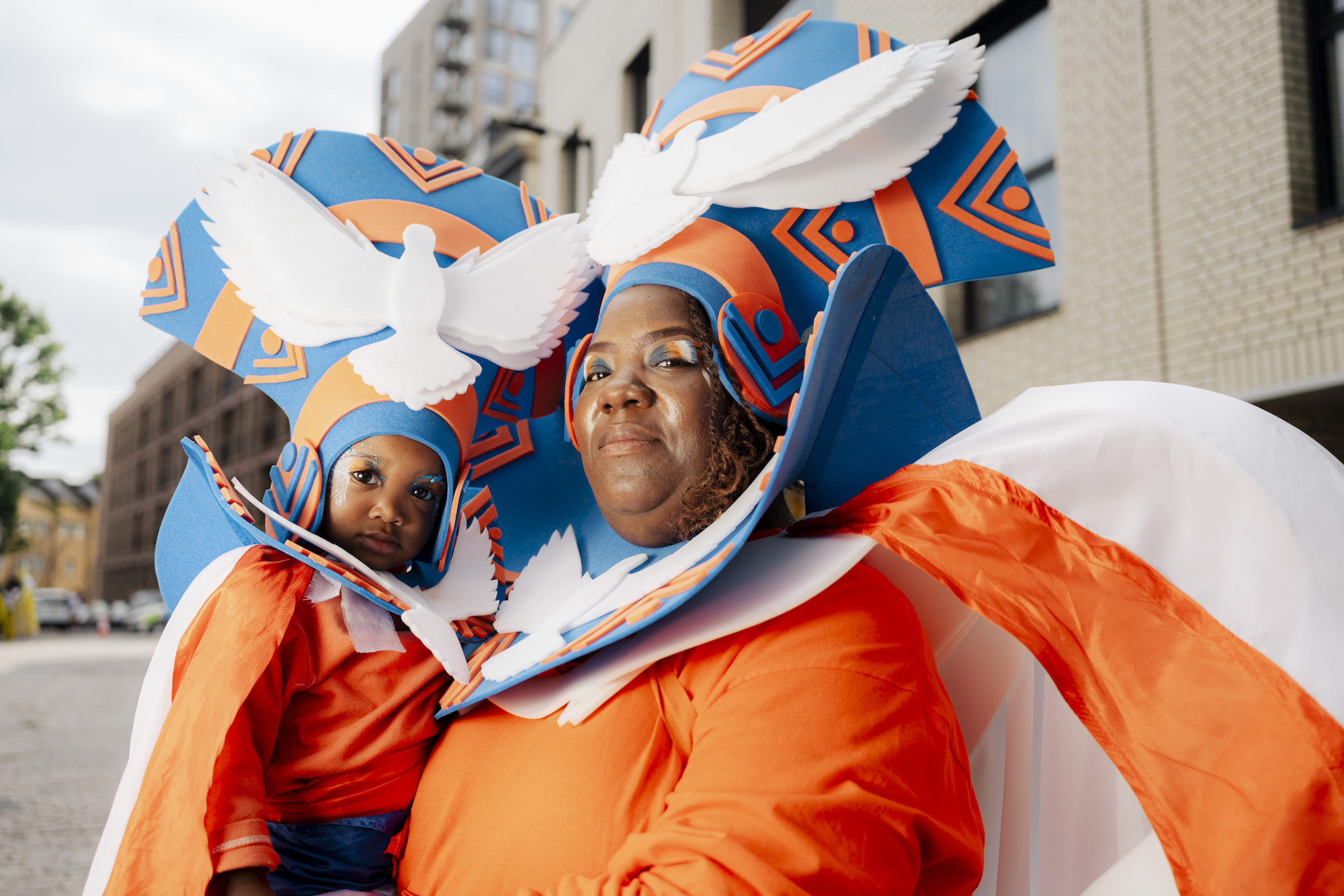 Two carnival performers wearing orange and white costumes and blue and orange headdresses, each with doves with outspread wings on them.