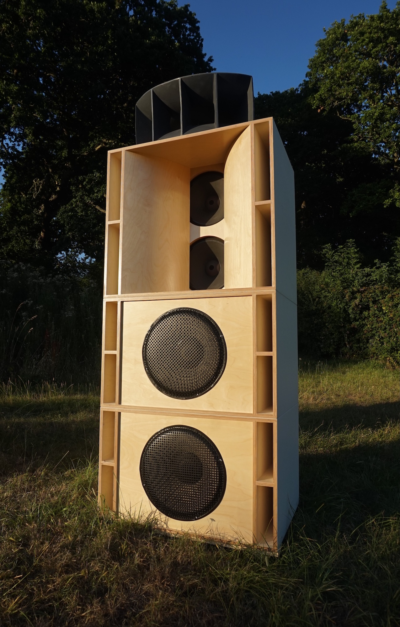 A tall wooden loudspeaker cabinet with two large circular drivers and horn openings at the top, standing on grass with trees and a blue sky behind it.