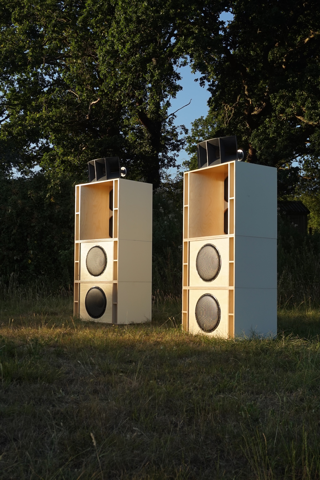 Two tall white outdoor speakers stand on grass among trees, each with three visible drivers and small units mounted on top.