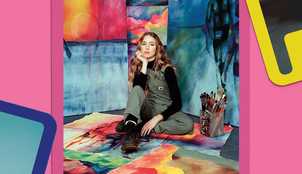 Woman with brown hair in green overalls and black top sitting cross-legged in colourful art studio with pink walls and rainbow floor.