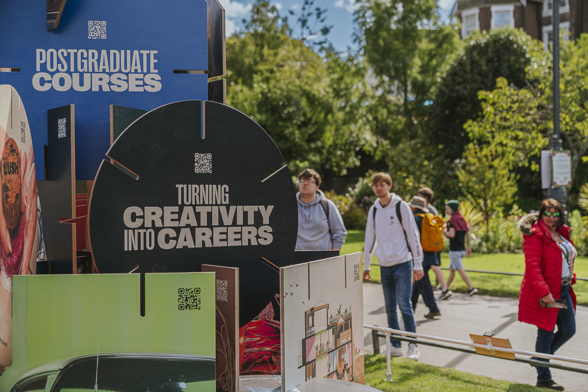 Close-up of a sculpture illustration based on Arts University Bournemouth's House of Cards prospectus in Bournemouth Gardens, focusing on a blue square card with text reading "Postgraduate Courses" and a black circle card with text reading "Turning Creativity into Careers".