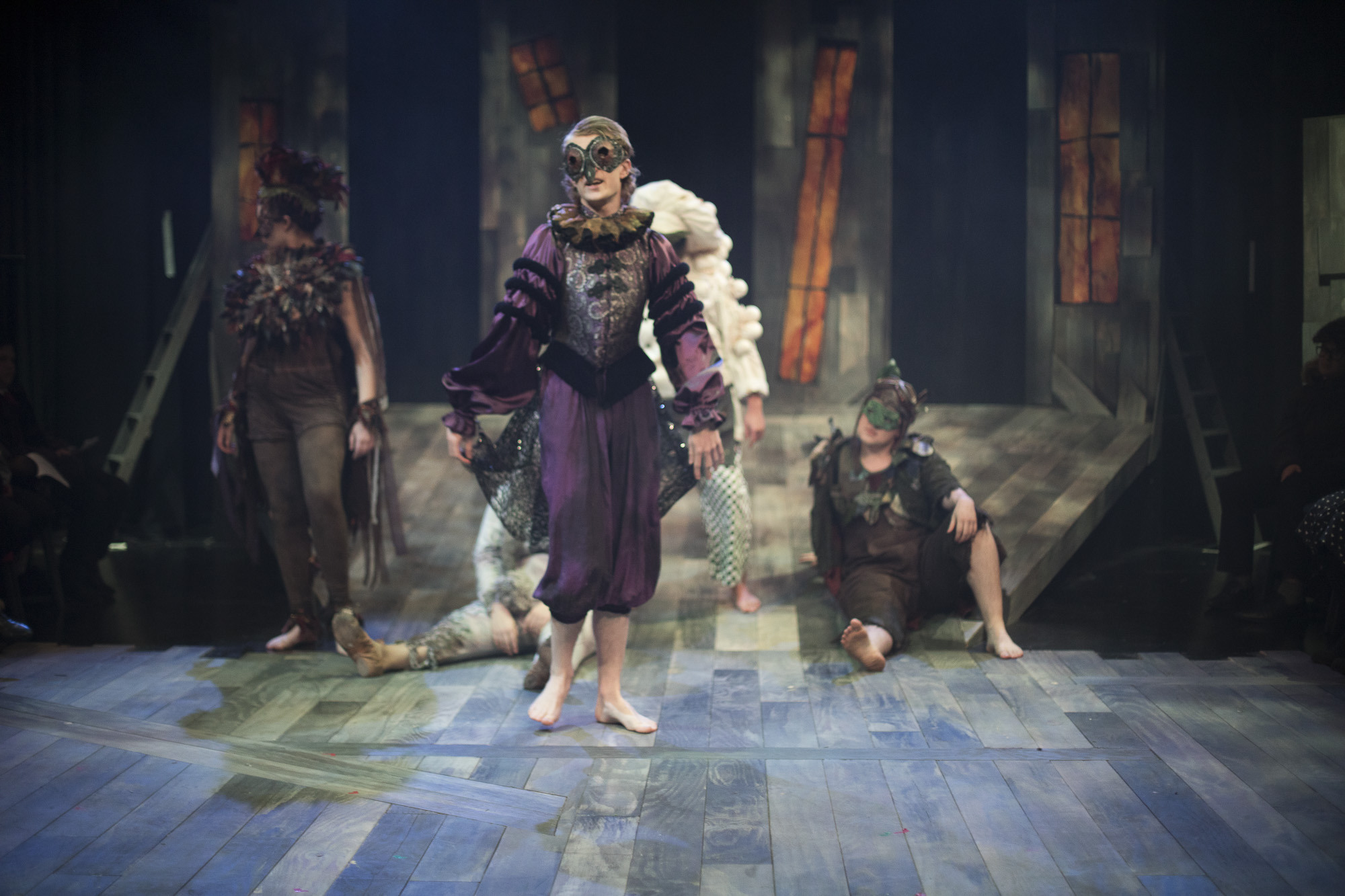 A performer in Venetian-style costume with a ruff and mask walks barefoot on a stage. Three other performers are behind them.