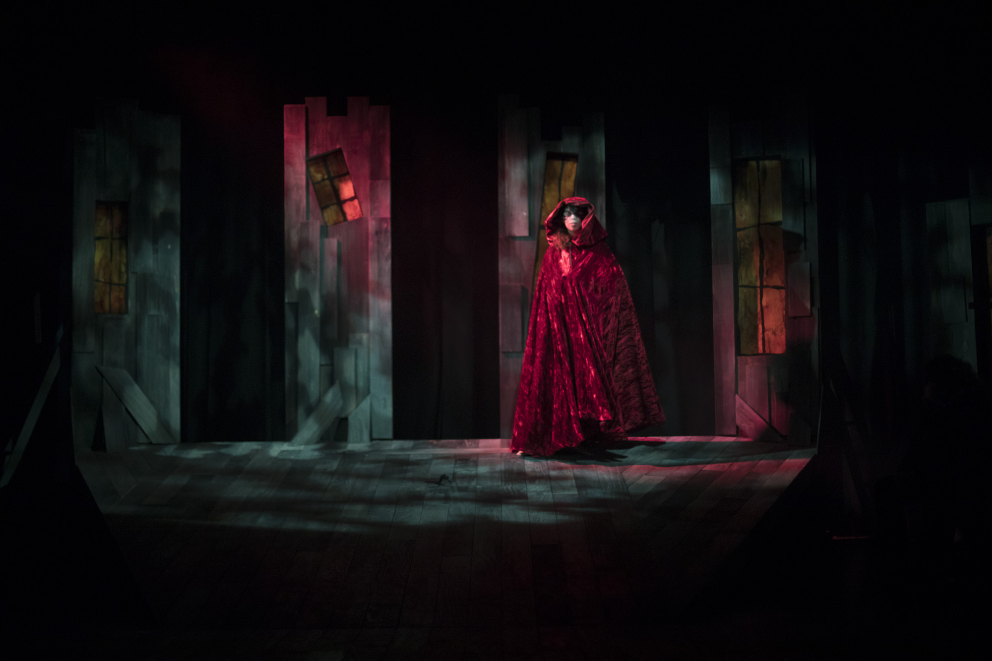 A person in a red hood and cloak stands on a stage in dim light with windowed panels behind them.