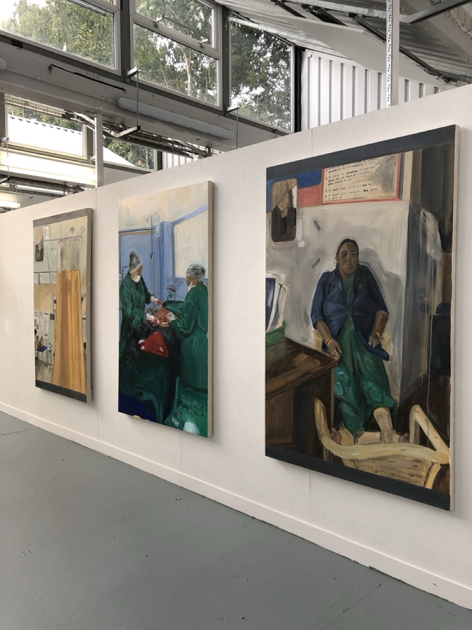 Three framed paintings on white gallery wall in industrial space with exposed ceiling and windows. Works feature figures in green, blue, and earth tones.