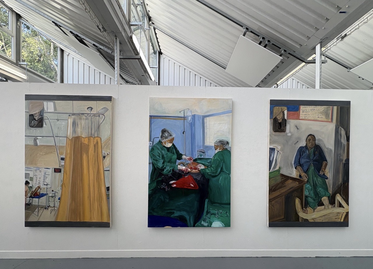 Gallery wall with three framed paintings: left shows yellow figure in white interior, centre depicts green-clad figures around red table, right shows blue-robed figure in brown setting.