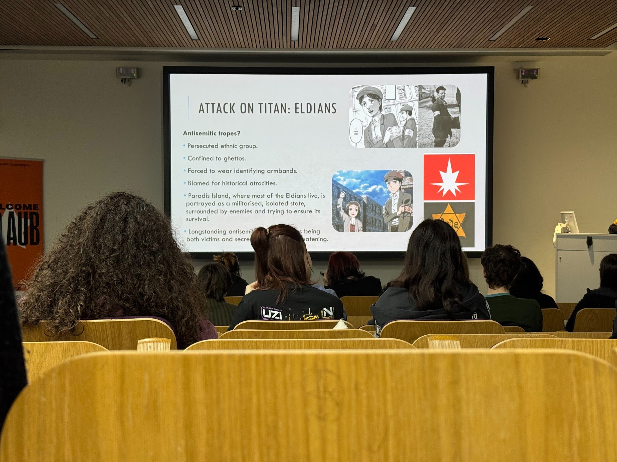 A presentation slide titled 'Attack on Titan: Eldians' displayed on a screen in a lecture hall. The slide features text outlining antisemitic themes and symbols, alongside images related to the topic. Attendees are seated in wooden chairs, viewed from behind.