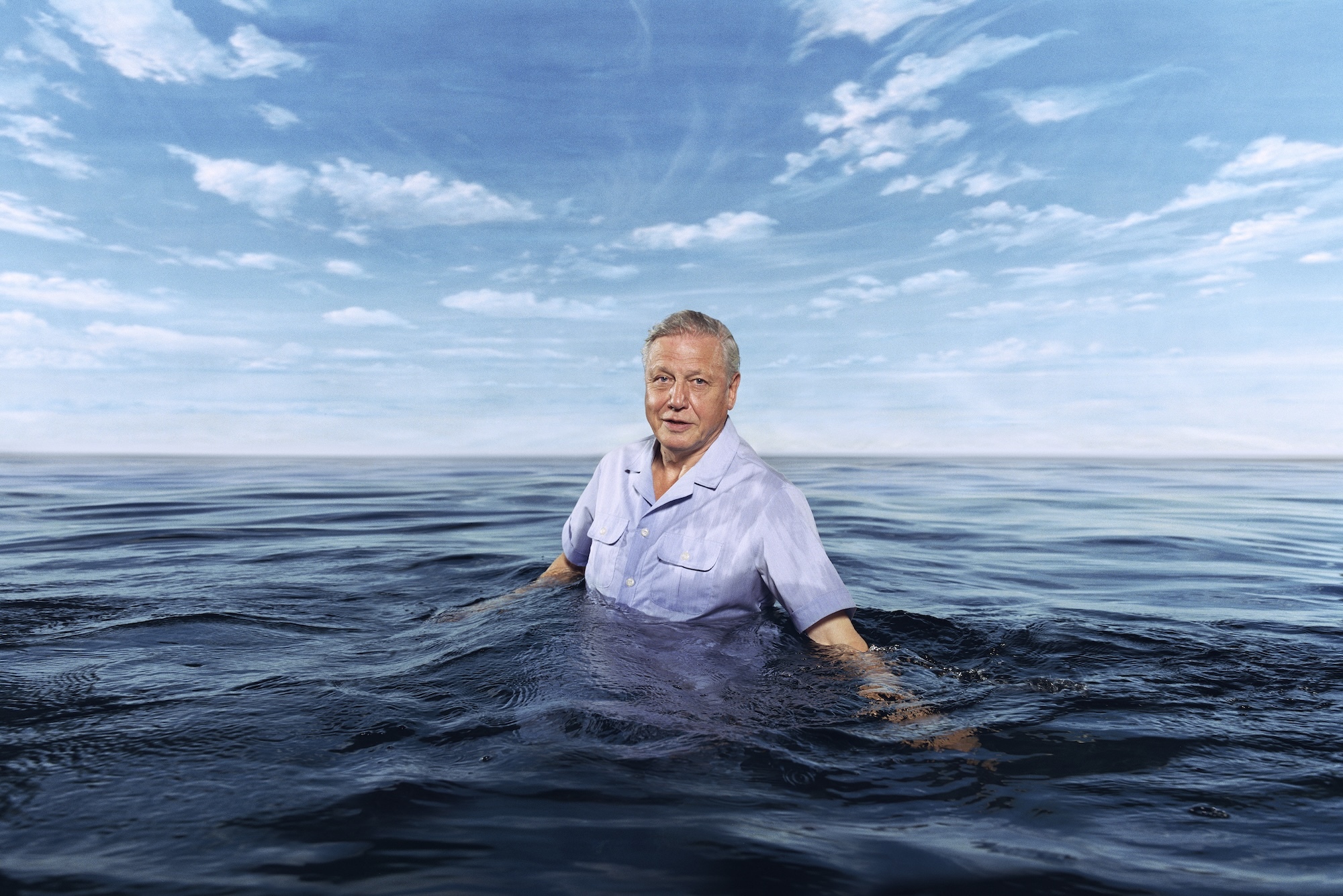 Sir David Attenborough standing waist-deep in dark blue ocean water under cloudy blue sky.