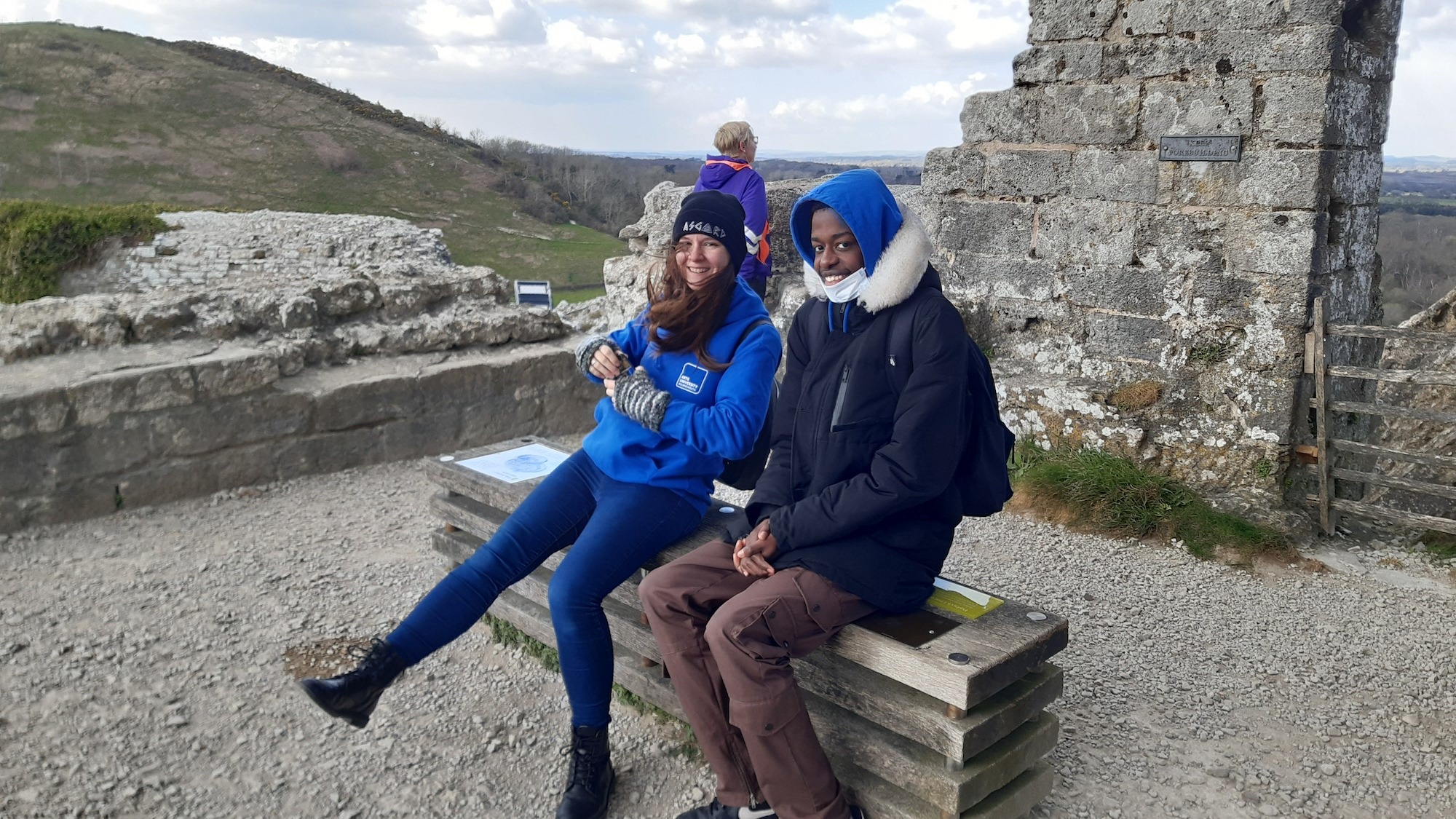 Two students sat on a bench at some historical ruins on a hill in cold weather.