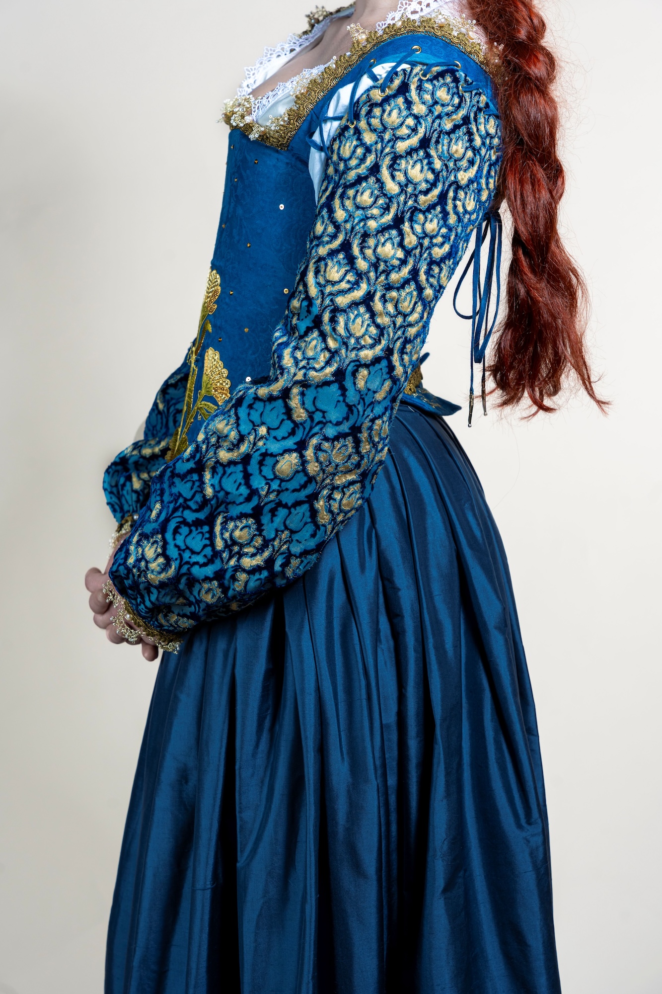 Young person viewed from the side wearing an ornate Renaissance blue gown with ornate patterned sleeves.