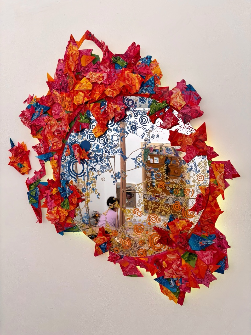 A circular piece of work on the wall consisting of a central mirror and colourful shapes and colours exuding around the edges.