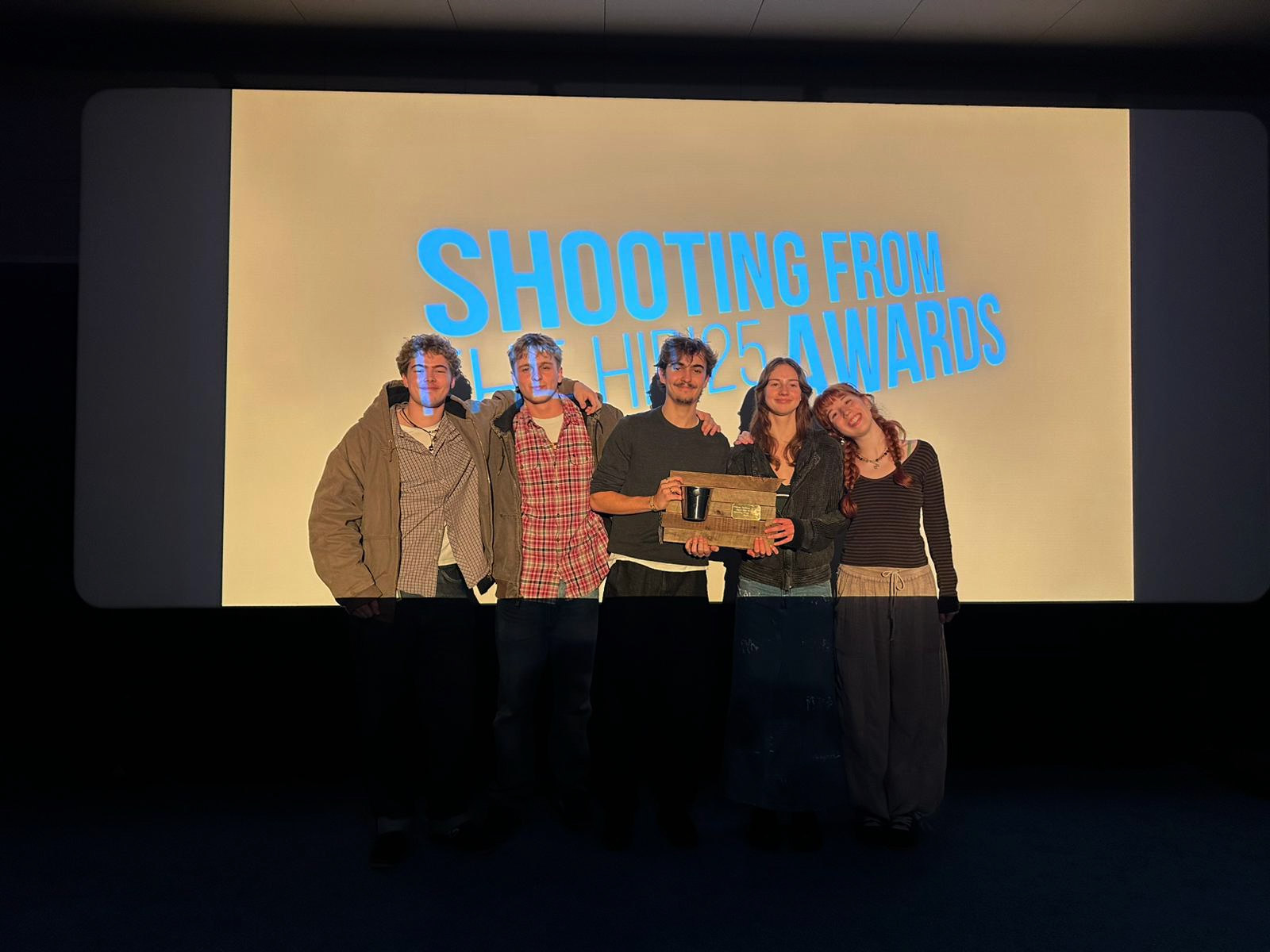 Five people standing on stage holding award, large screen behind displays "SHOOTING AWARDS" in blue text on cream background.
