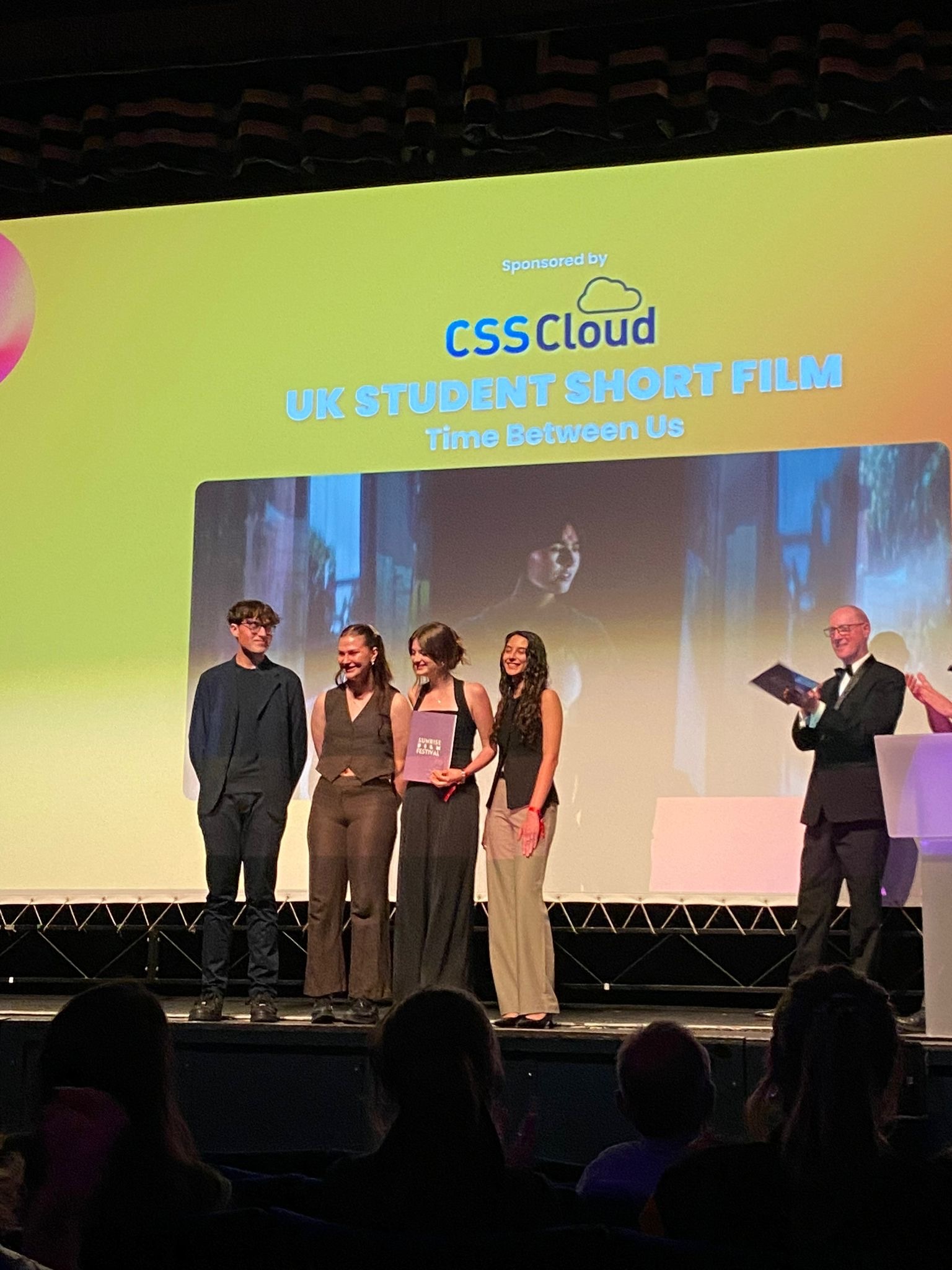 Five people on stage at awards ceremony, man in suit presenting to four recipients, large screen showing "CSSCloud VR Student Short Film" award.