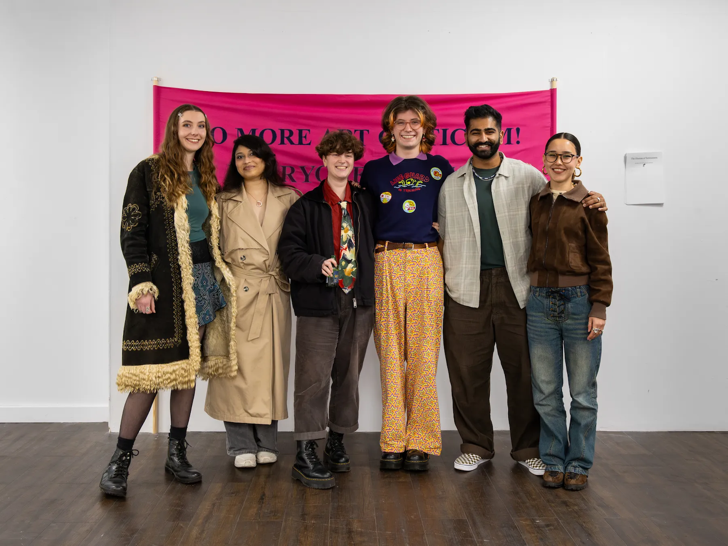 Six people standing together in front of a bright pink banner on a white gallery wall, with dark wooden flooring beneath them.