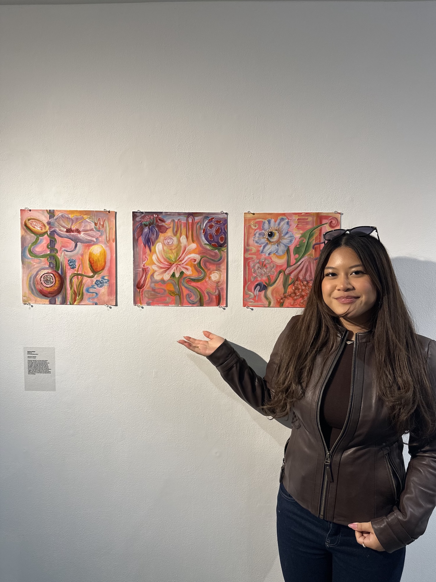 Illustrator A. Shukor stands next to three colourful abstract paintings displayed on a white wall. She is pointing towards the paintings, while a small label is visible on the wall beneath them.