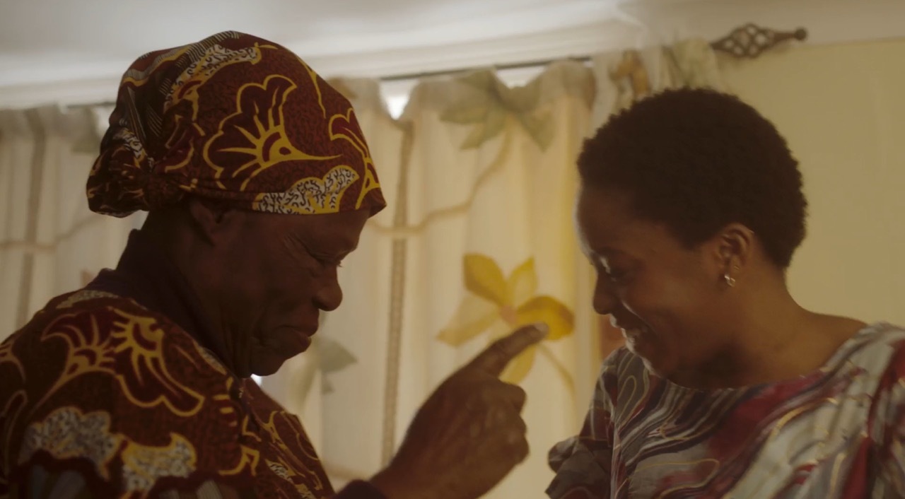 Two women indoors, one wearing patterned headwrap and clothing, the other in light-coloured top, both looking down between them.