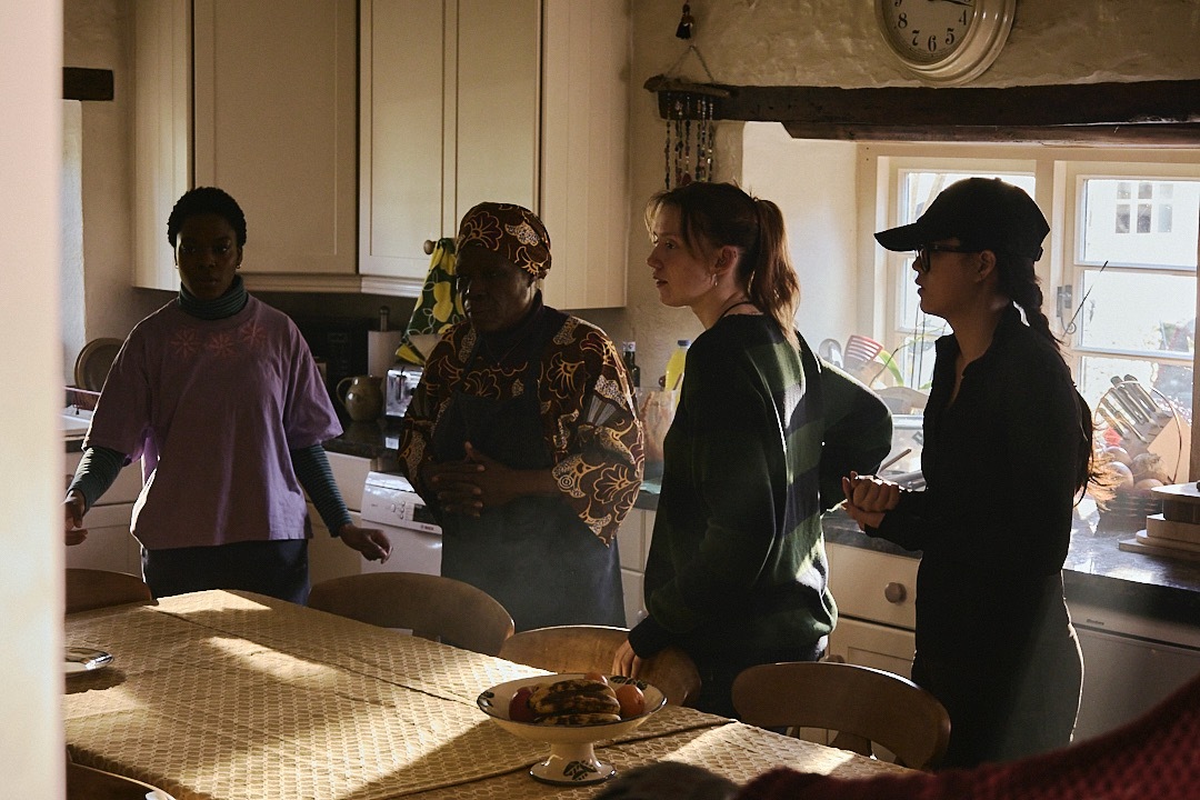 Four people standing in a kitchen with cream cabinets, one wearing a patterned top, another in dark clothing with a cap.