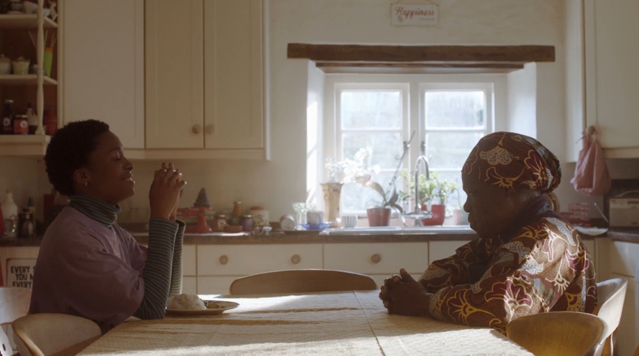 Two people sitting at kitchen table, one drinking from green bottle. White cabinets, window with plants on sill, warm natural lighting.