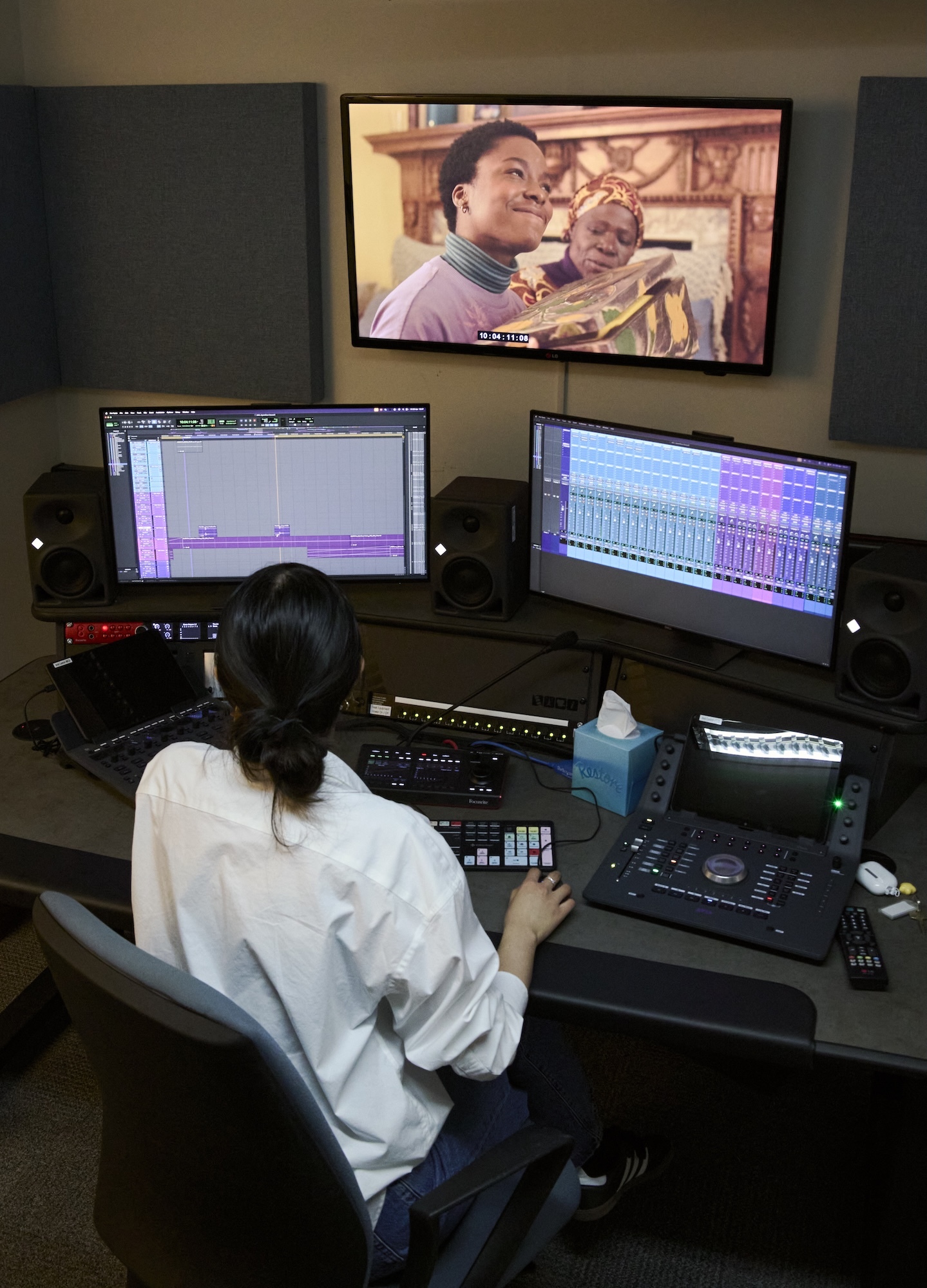 A BA (Hons) Film Production student in white shirt working at audio editing station with two computer monitors and mixing board, television screen above showing programme.