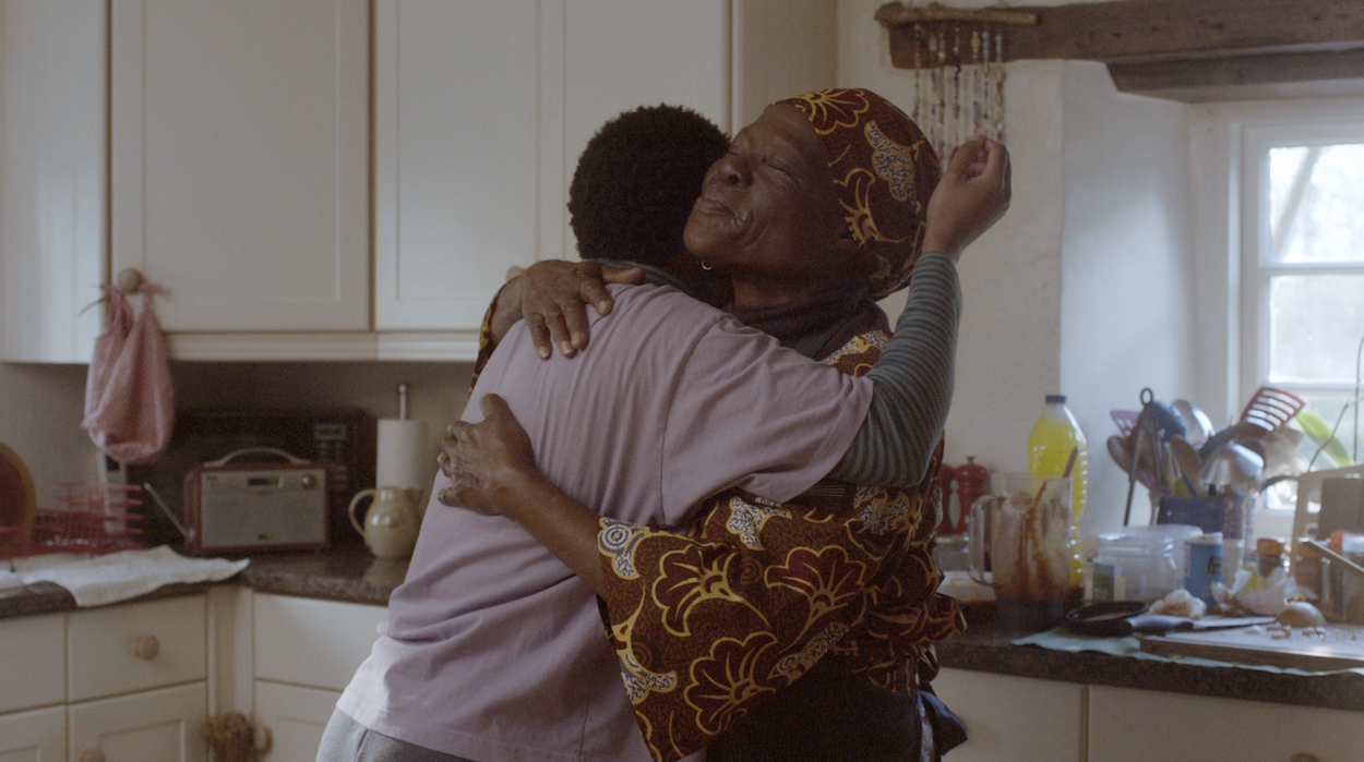 Still from the film "Ayomikun". Two people embracing in a kitchen with white cabinets, one wearing a patterned dress and head scarf, the other in light clothing.