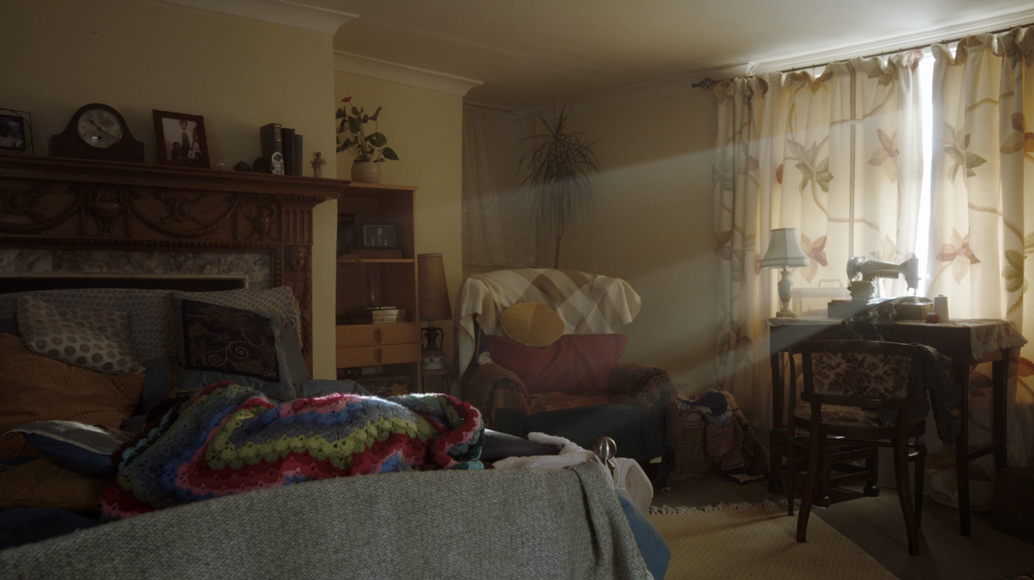 Still from the film "Ayomikun". Living room with beige sofa, dark wooden furniture, patterned curtains, and colourful blankets on grey chair in warm lighting.
