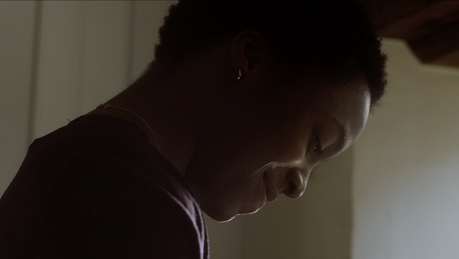 Still from the film "Ayomikun". Actor with dark curly hair looking down, wearing dark clothing, dimly lit with shadows across face and warm background lighting.
