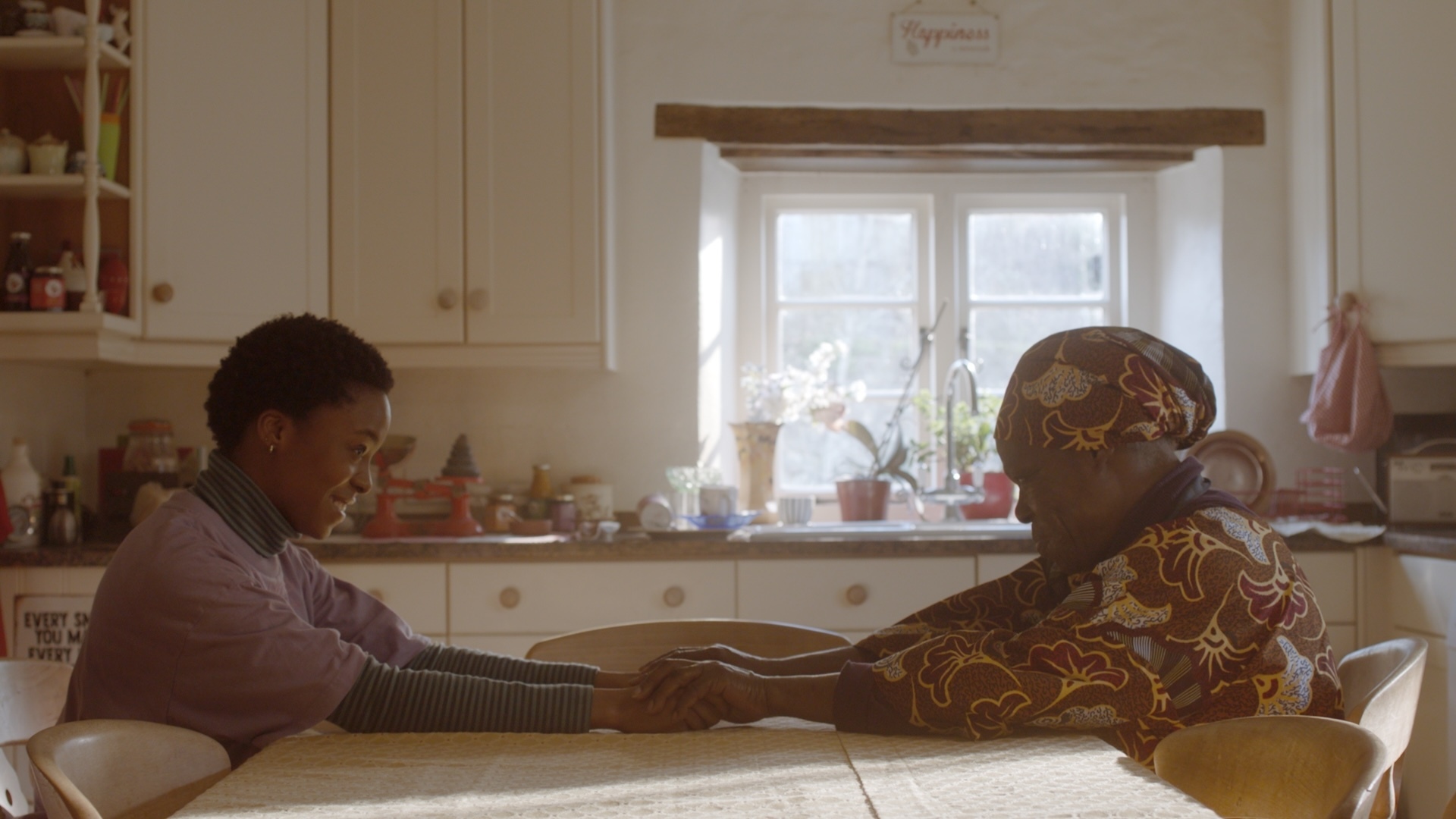 Still from the film "Ayomikun". Two people sitting at wooden kitchen table, one in purple jumper, other in patterned yellow clothing with matching headwrap. White cabinets, window behind.