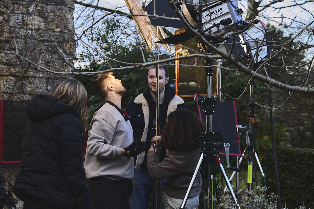 Two BA (Hons) Film Production students operating film equipment on tripod beneath bare tree branches with buildings visible in background.