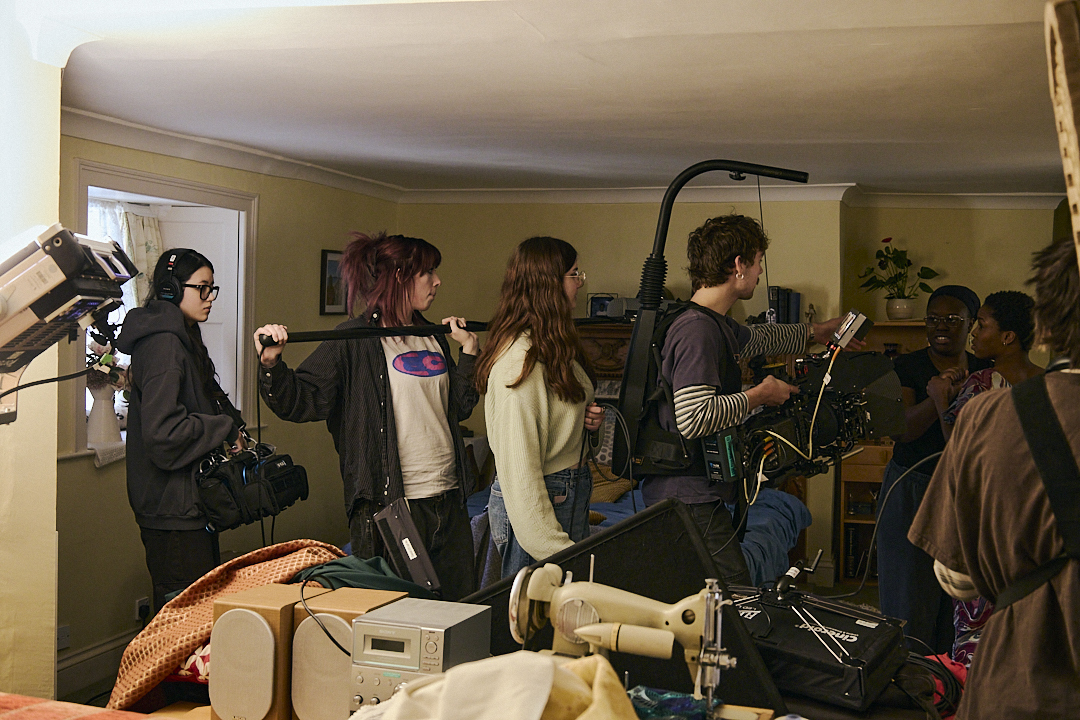 Film crew with cameras, lighting equipment, and microphones crowded in small beige room during production shoot.