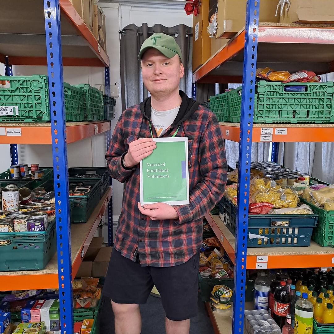 Ollie Wheaton holding a green food bank writing anthology in a food bank, stacks of green crates on shelves.