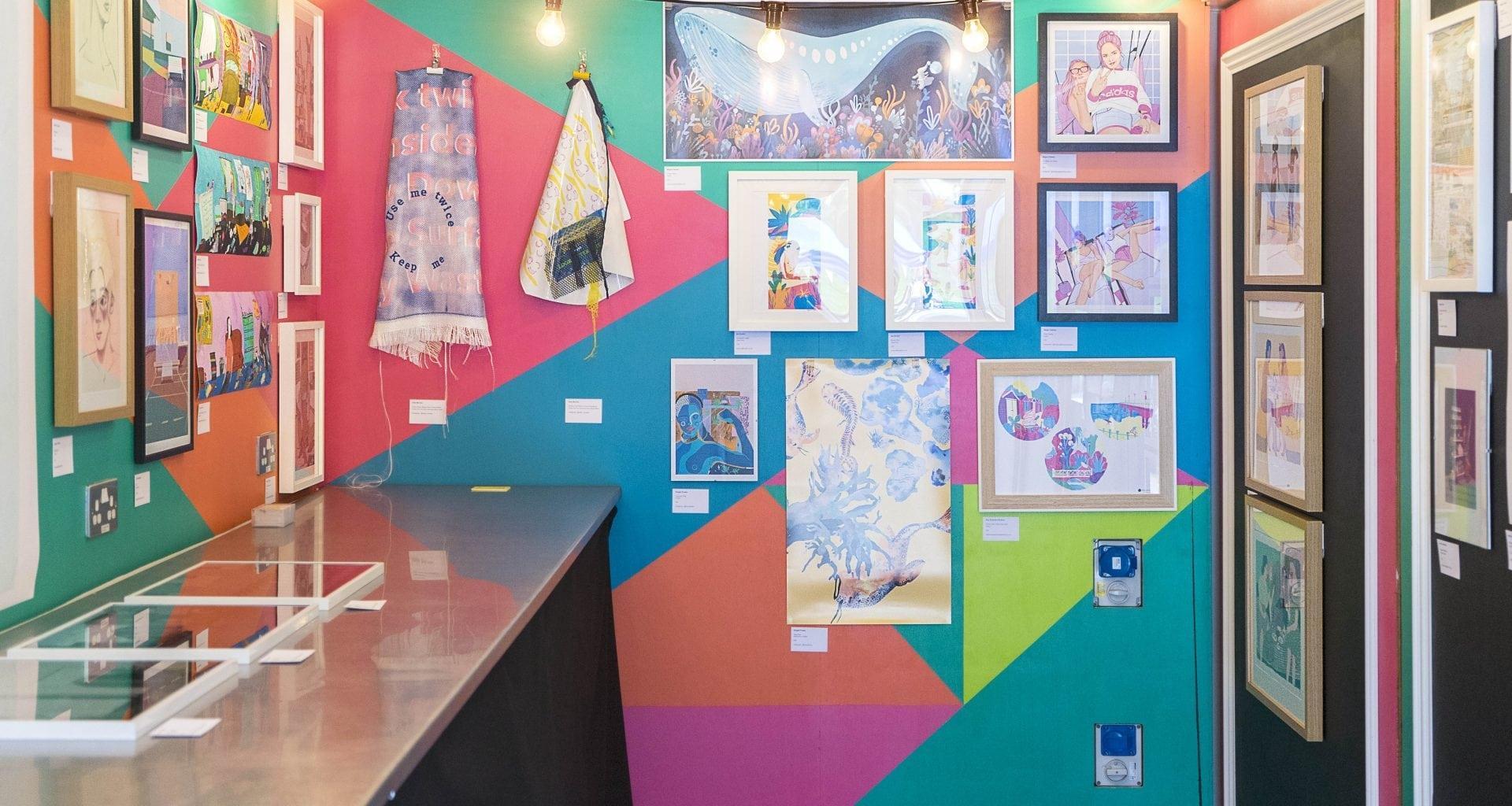 Three walls of a gallery space painted in bright colours. A collection of exhibits including textiles and framed artworks are displayed on the walls.