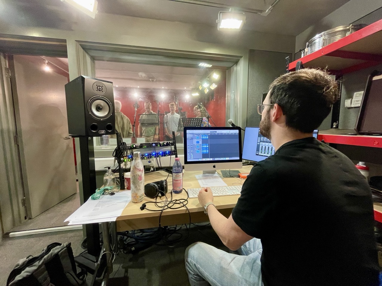 Man in black shirt working at desk with computer monitors, studio monitor speaker, and audio equipment in recording studio.