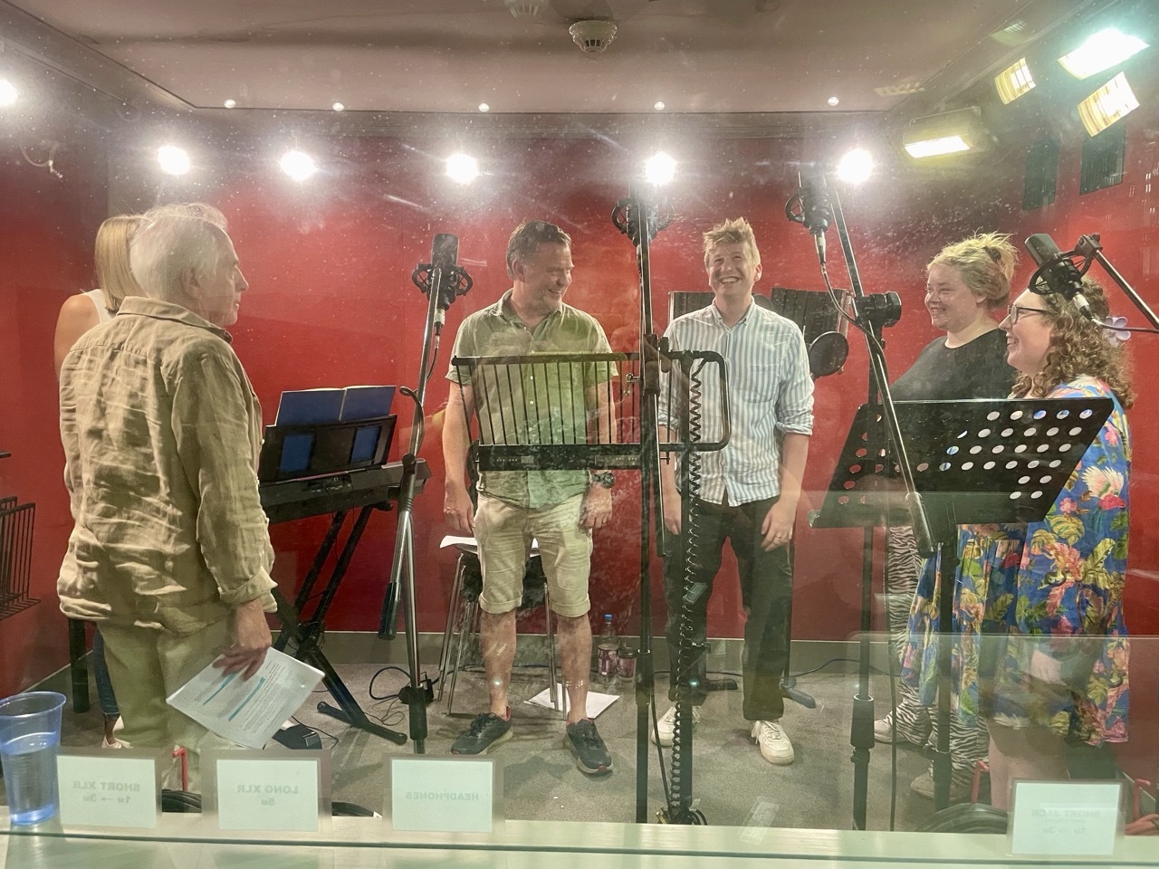 Four people in recording studio with microphones, music stands, and red walls. Glass partition separates performers from control room.