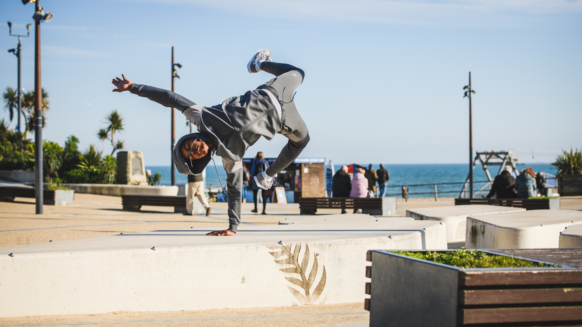 BA (Hons) Dance student posing in the air, standing on one hand, at Bournemouth Pier Approach, with Bournemouth Beach and the sea in the background.