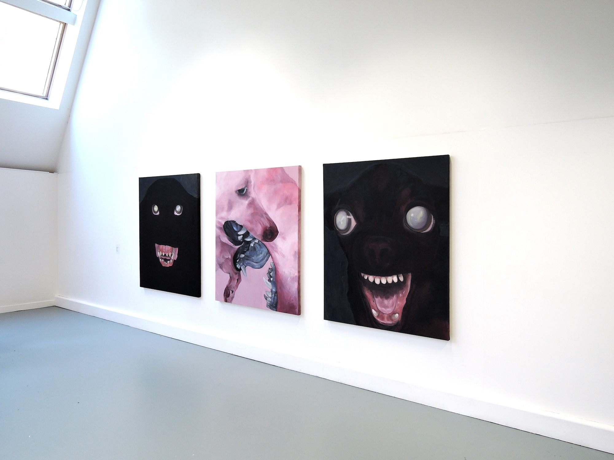 Three artworks mounted on white gallery wall: left black canvas with small figures, centre pink/purple abstract piece, right black canvas with white circular shapes and red form below.