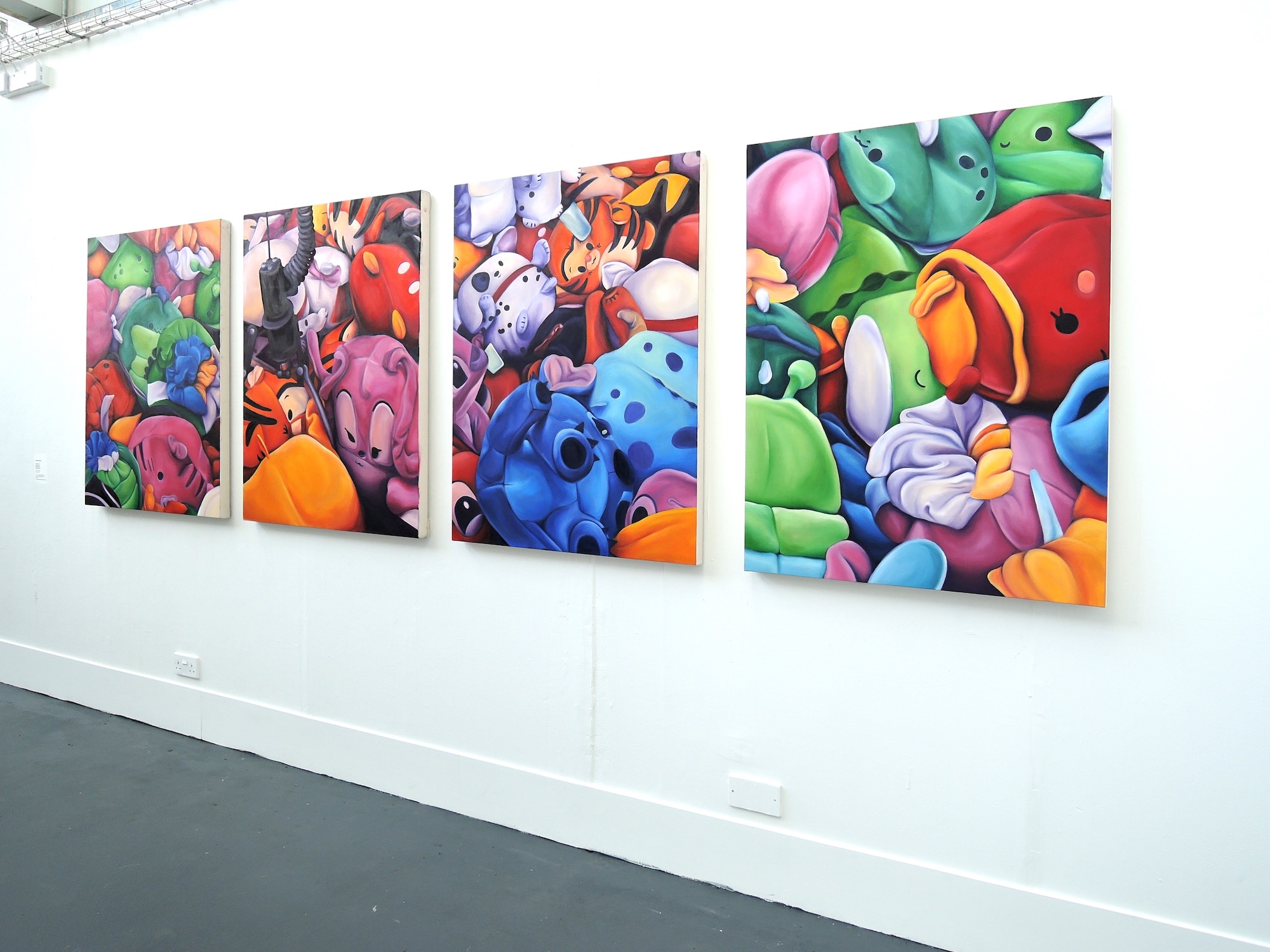 Three large colourful paintings on white gallery wall featuring bright geometric shapes and cartoon-like forms in vibrant blues, greens, reds, and oranges.