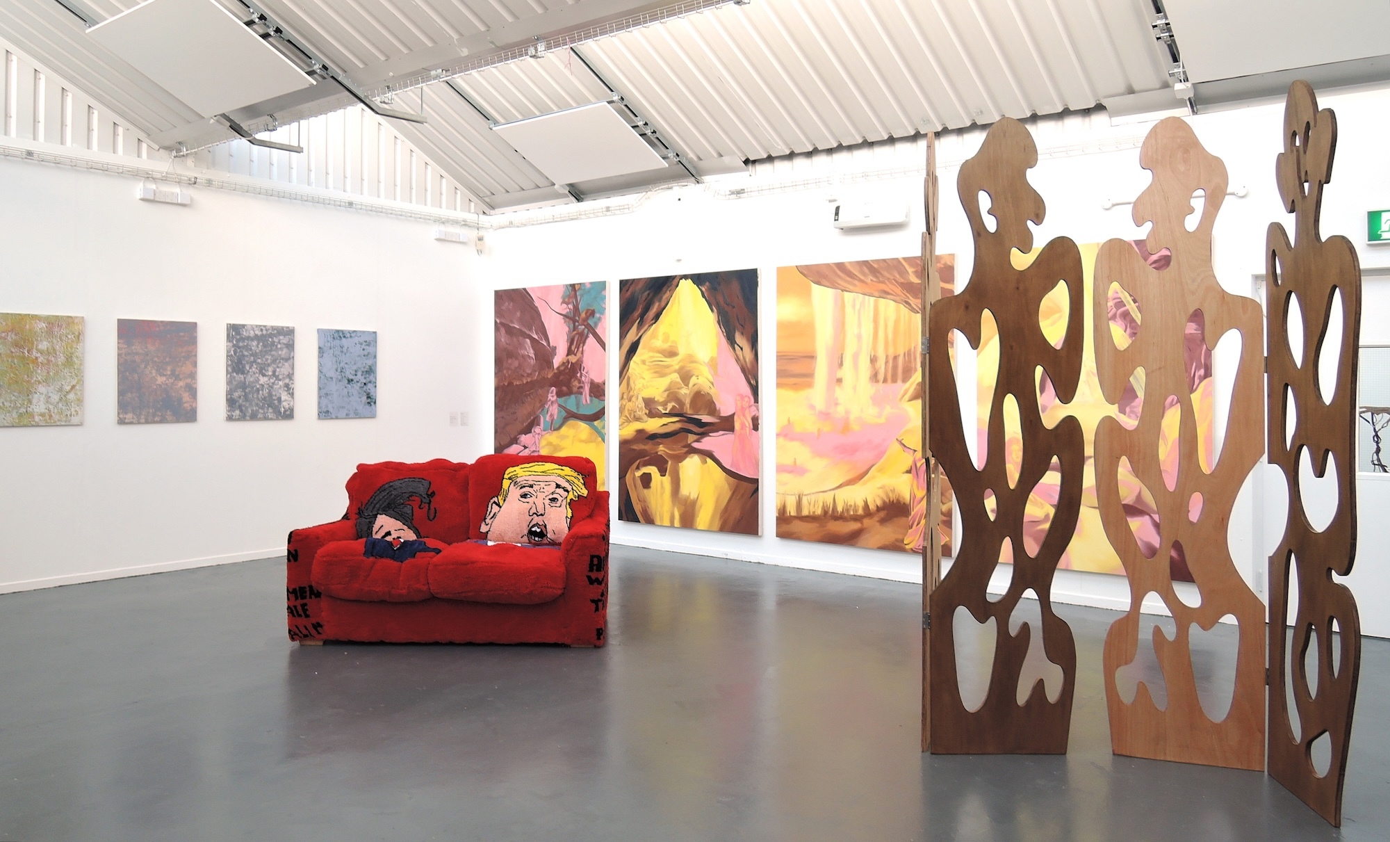 Gallery space with white walls displaying colourful artwork, red sculptural sofa, and brown perforated room divider panels.