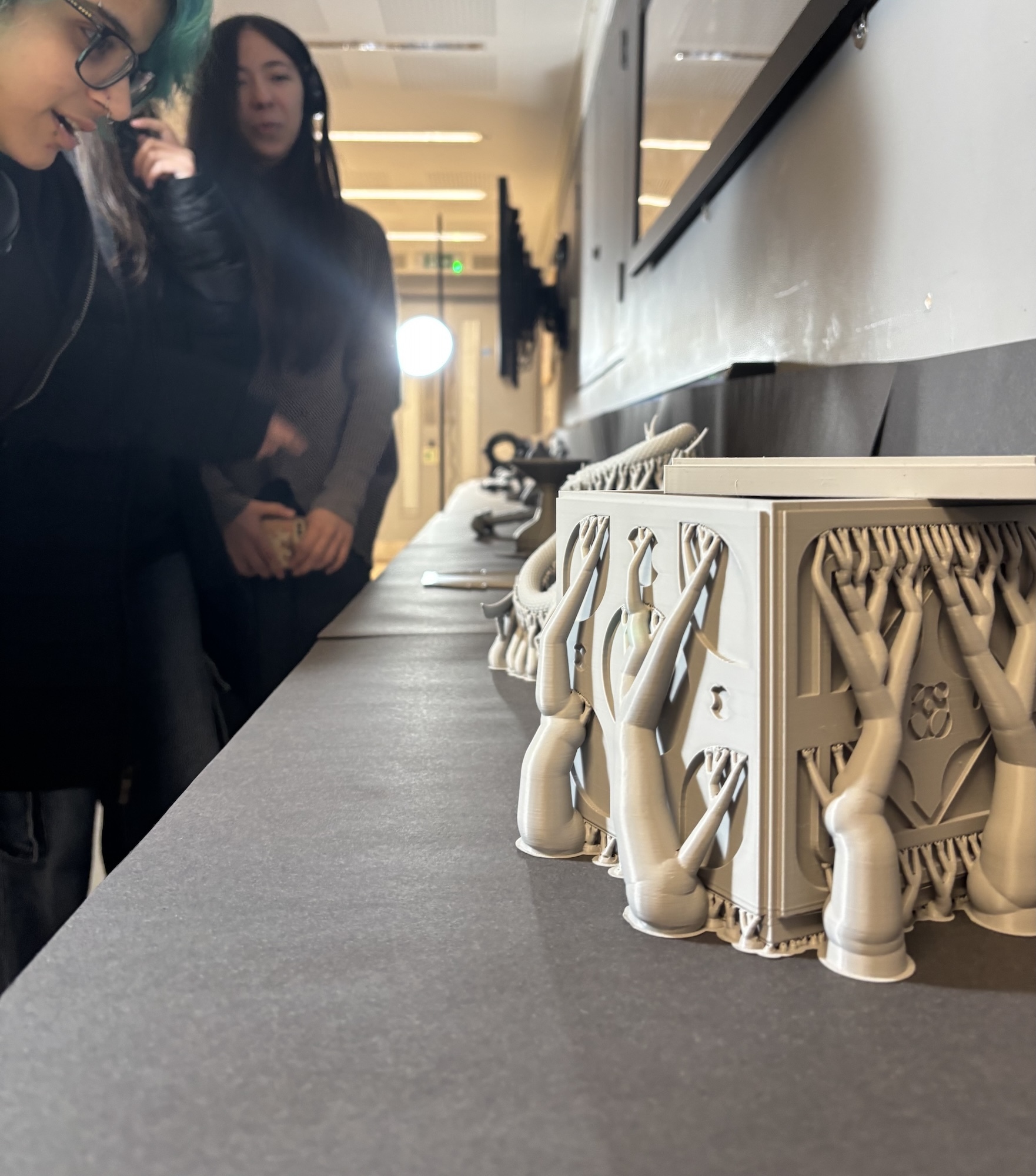 Two women examining white ceramic or plaster sculptural objects displayed on shelves in a bright gallery space with white walls.