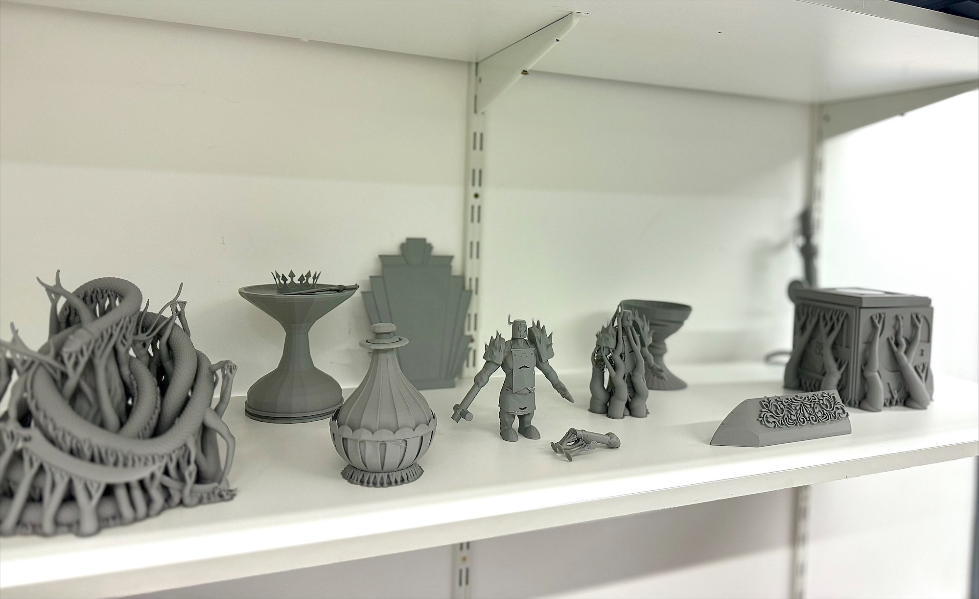 Grey 3D printed objects with intricate geometric patterns and spiky textures displayed on white shelving unit against pale wall.