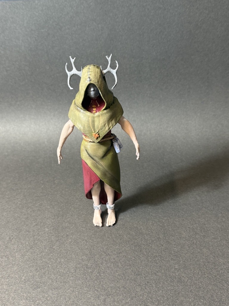 Action figure with green hooded robe, white antlers, red garment underneath, pale skin, standing on grey surface against grey backdrop.