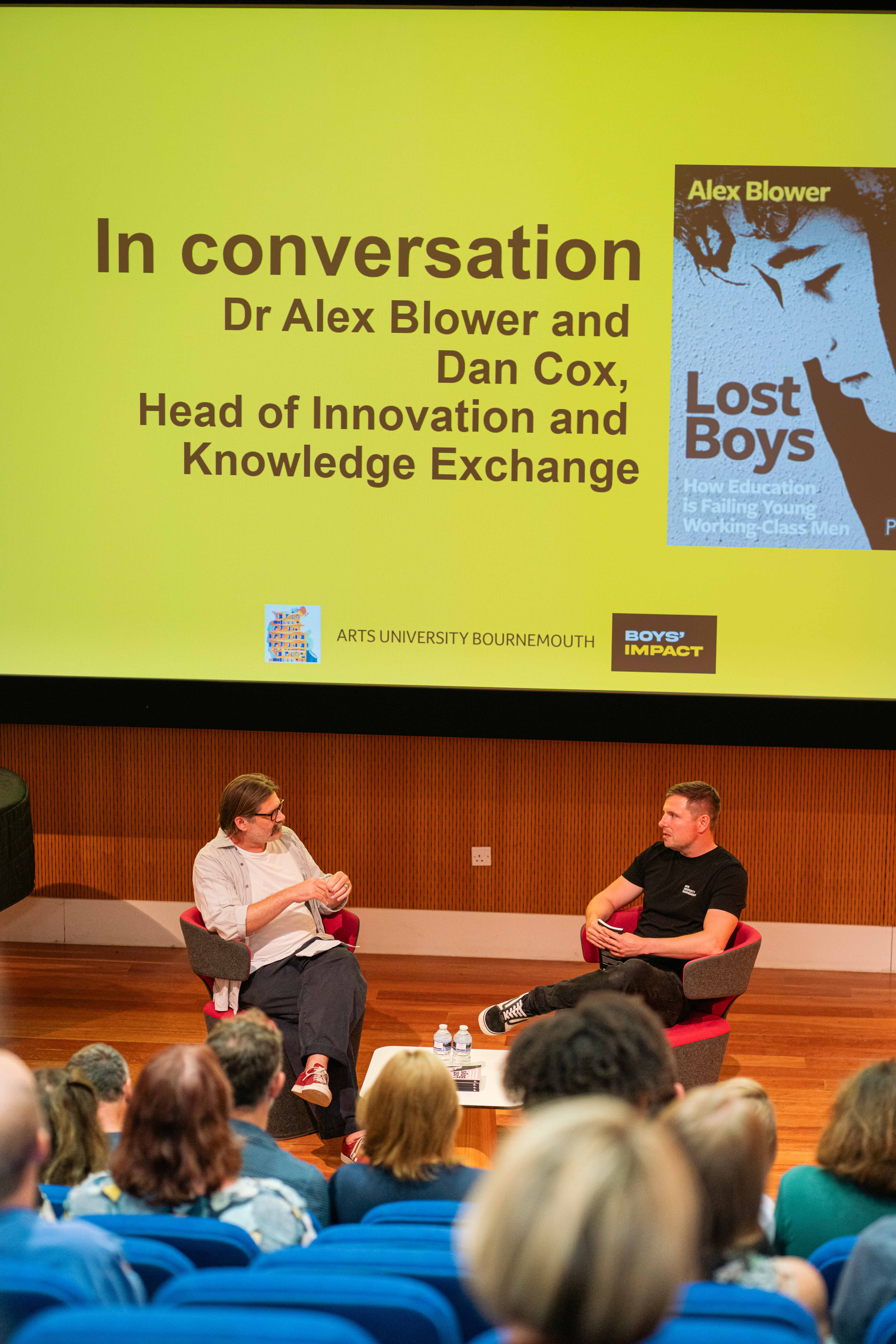 D Cox and Dr A. Blower seated in red chairs on stage speaking to audience, with yellow projection screen displaying "In conversation" text and book cover.