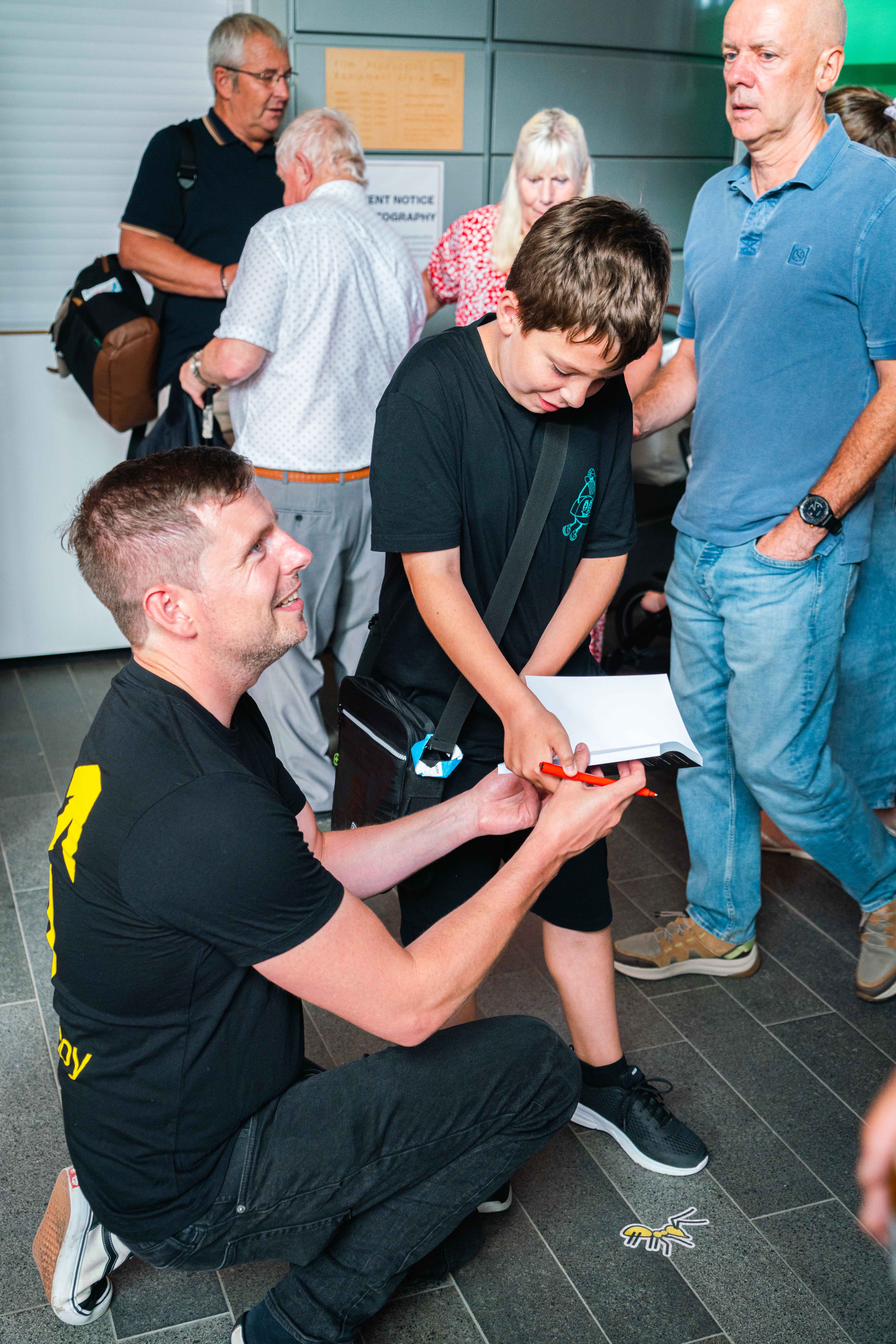 Dr A. Blower kneeling whilst signing a copy of his book for a young person, surrounded by adults watching in indoor setting.
