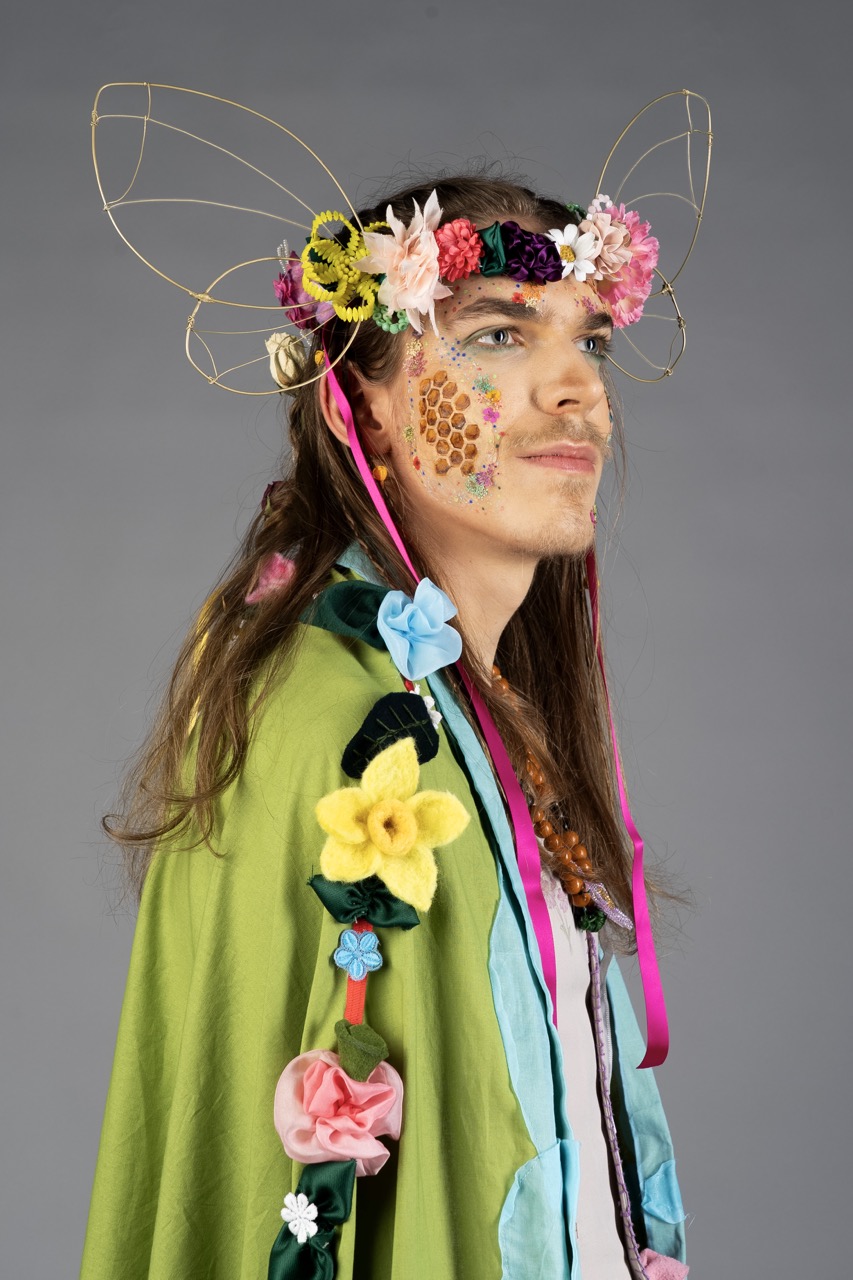 A person with long hair wearing a floral headpiece and wire fairy wings, dressed in a green and blue costume against a grey background.