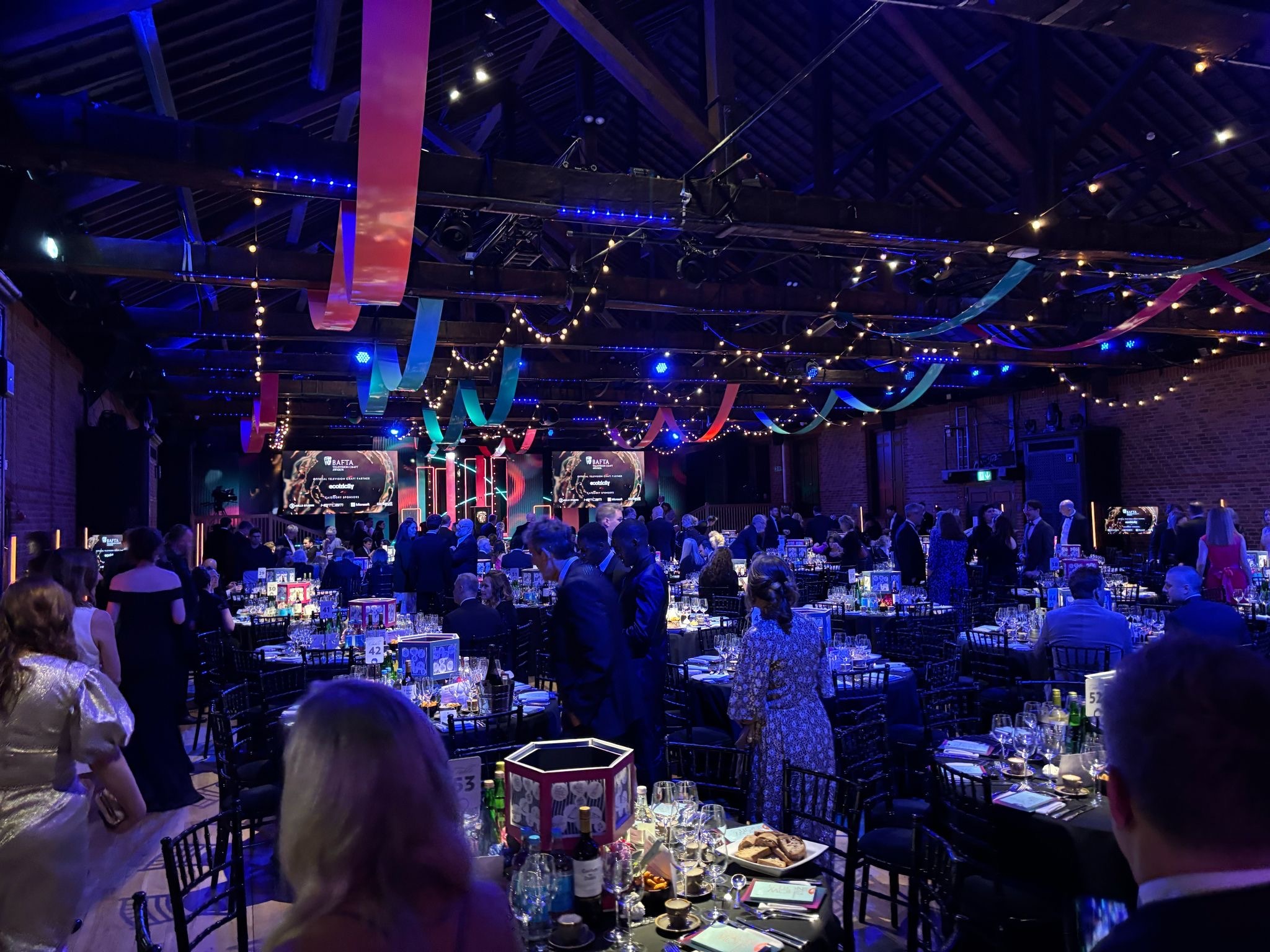 Large event space with blue and purple lighting, exposed ceiling, round tables with guests seated, and decorative hanging lights.